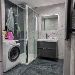 Mateusz Wardyński - Nowoczesna łazienka po remoncie w Świnoujściu: kabina prysznicowa, umywalka z szafką, pralka. Marmurowe płytki na ścianach i podłodze. Oświetlone lustro.