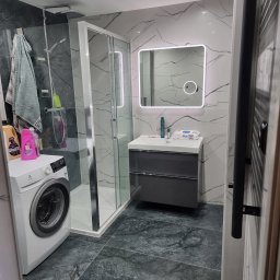 Mateusz Wardyński - Nowoczesna łazienka po remoncie: kabina prysznicowa, umywalka z szafką, pralka i marmurowe płytki na ścianach i podłodze. Całość utrzymana w szaro-białej kolorystyce.