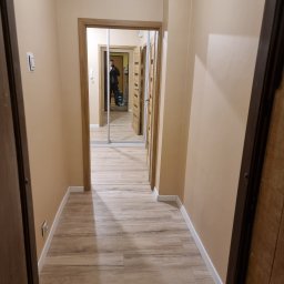 Mateusz Wardyński - Beżowy korytarz z drewnopodobną podłogą i lustrem w zabudowie. Widoczna osoba robiąca zdjęcie. Minimalistyczne wykończenie, ciepłe oświetlenie.