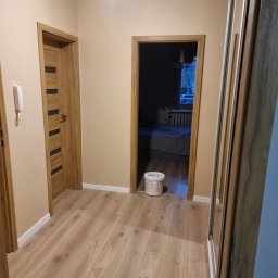 Mateusz Wardyński - Beżowy korytarz po remoncie z drewnianą podłogą, dwojgiem drzwi i białą puszką farby. Widoczna szafa z lustrem i lampa sufitowa.