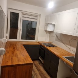 Mateusz Wardyński - Nowoczesna kuchnia w bloku: drewniany blat, szare szafki dolne, białe górne, marmurowe wykończenie ściany. Widok na okno i fragment podłogi z paneli.