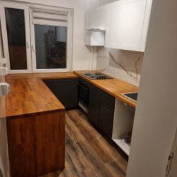 Mateusz Wardyński - Nowoczesna kuchnia po remoncie: drewniany blat, ciemne szafki dolne, białe górne, marmurowe wykończenie ściany nad blatem, drewnopodobna podłoga.