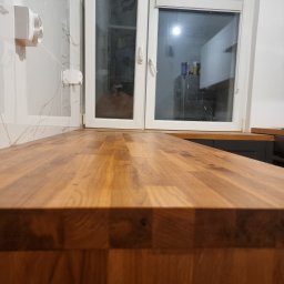 Mateusz Wardyński - Drewniany blat kuchenny z widocznym oknem i marmurową ścianą w tle. Ujęcie z niskiej perspektywy podkreśla strukturę drewna i nowoczesny design kuchni.