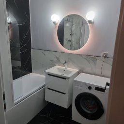 Mateusz Wardyński - Nowoczesna łazienka po remoncie: czarne płytki imitujące marmur, okrągłe lustro z podświetleniem LED, biała umywalka z szafką i pralka. Minimalistyczny design.