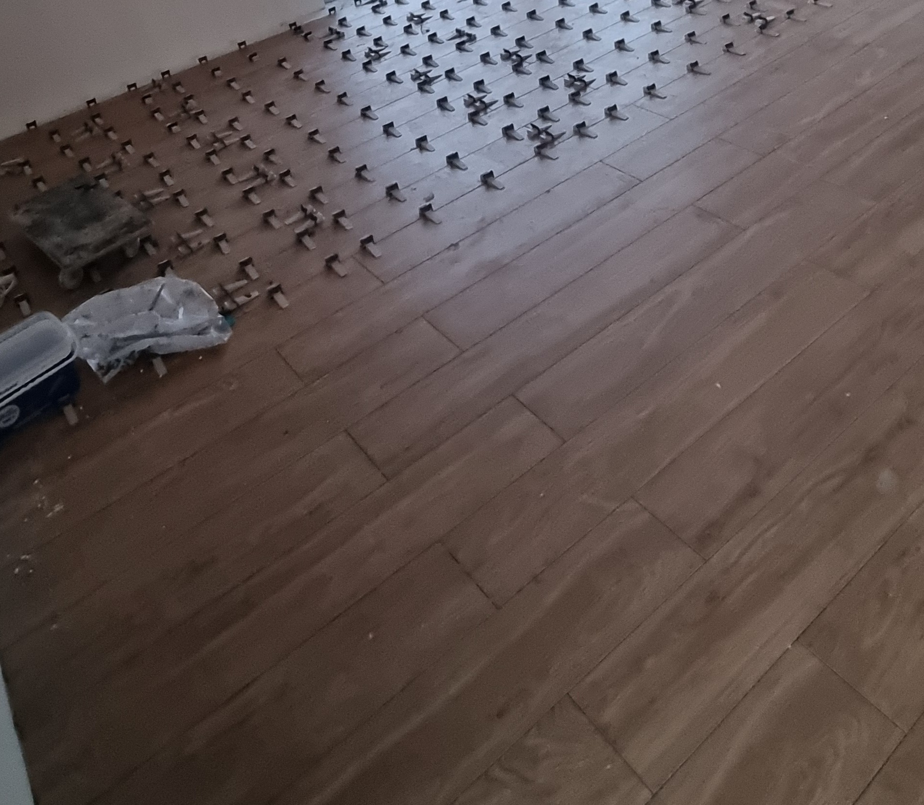 Układanie brązowych płytek podłogowych z systemem poziomowania. Widoczne narzędzia glazurnicze i fragment ściany. Ukończona część podłogi.