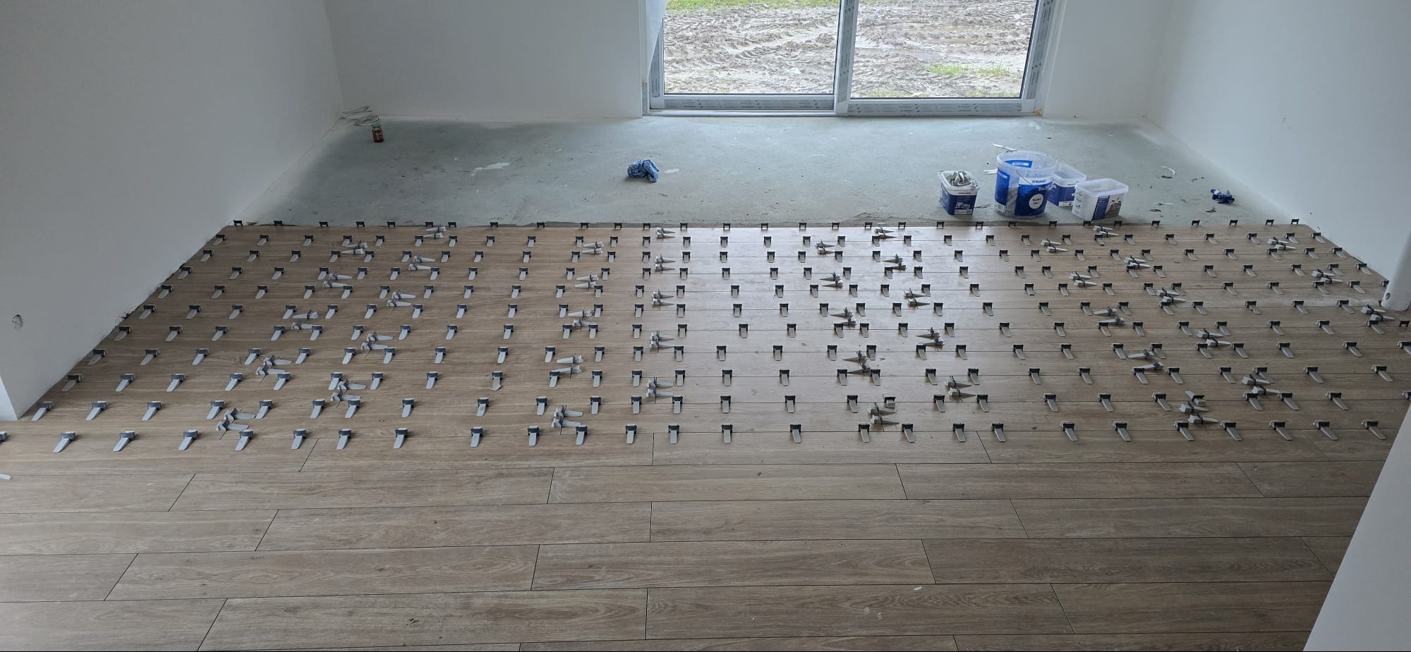 Układanie podłogi z płytek imitujących drewno, z użyciem systemu poziomowania płytek. Widoczne pojemniki z klejem i okno w tle.