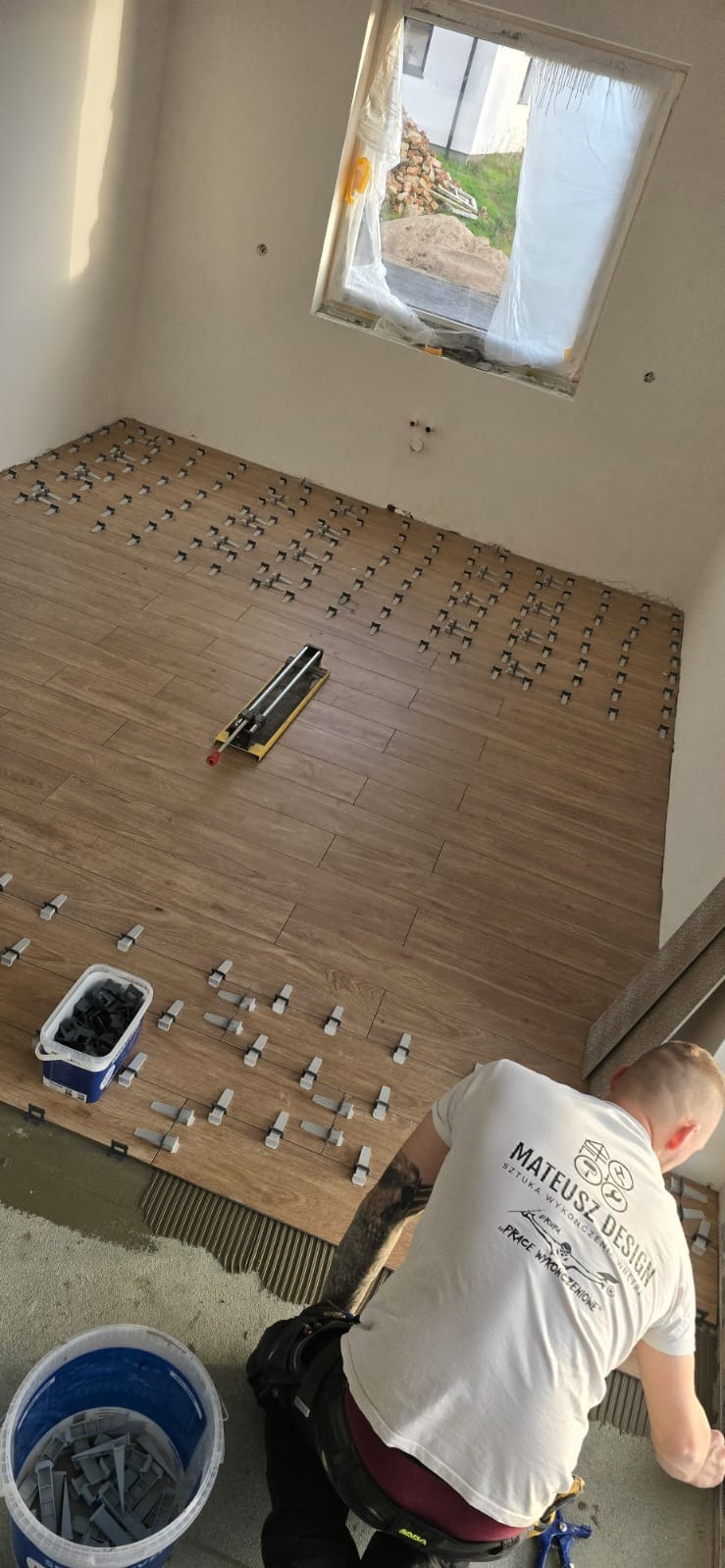 Układanie drewnopodobnych płytek podłogowych z użyciem klipsów poziomujących. Widoczny glazurnik w trakcie pracy, narzędzia i wiadro z klipsami.