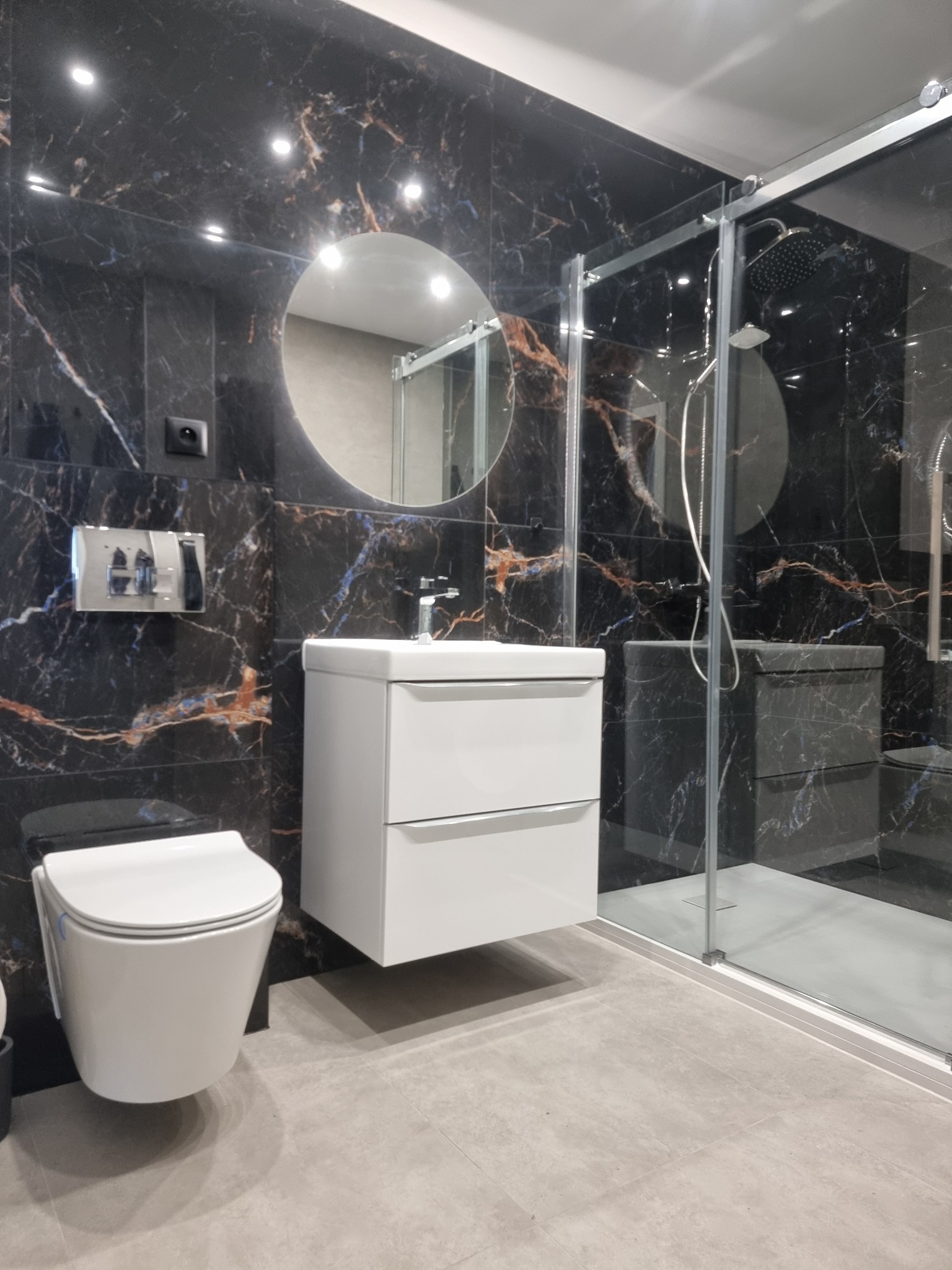 Nowoczesna łazienka z czarnymi płytkami imitującymi marmur, białą szafką z umywalką, okrągłym lustrem i kabiną prysznicową ze szklanymi drzwiami.