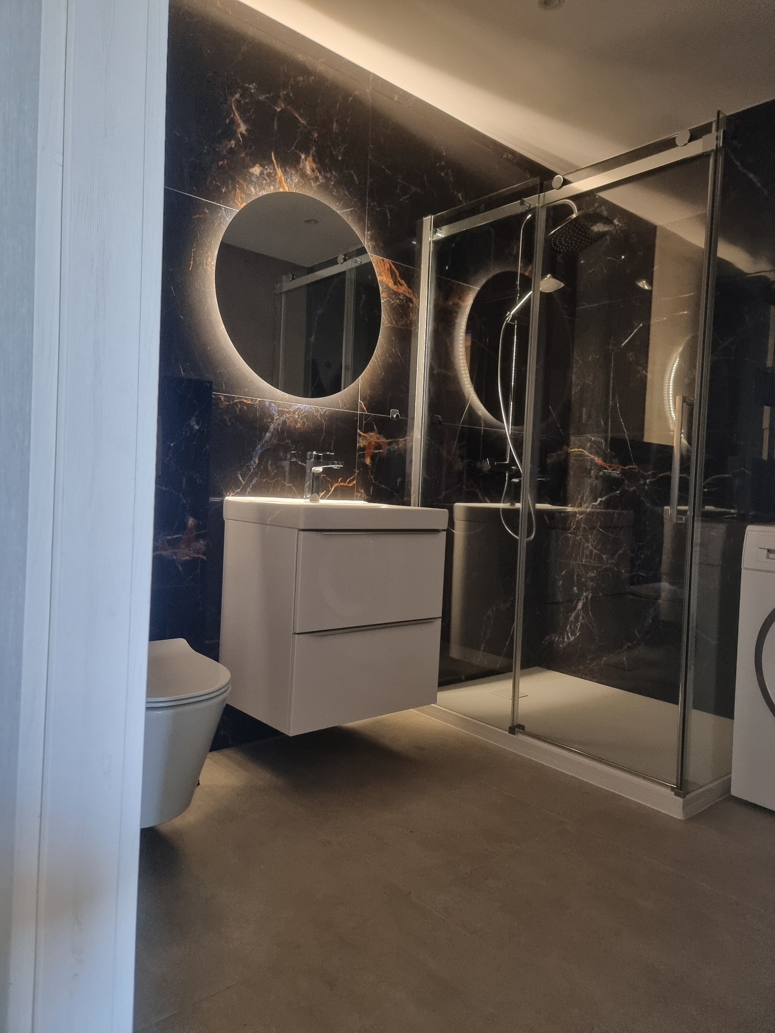 Nowoczesna łazienka z czarnymi płytkami imitującymi marmur, okrągłym lustrem z podświetleniem LED, białą szafką pod umywalką i kabiną prysznicową.