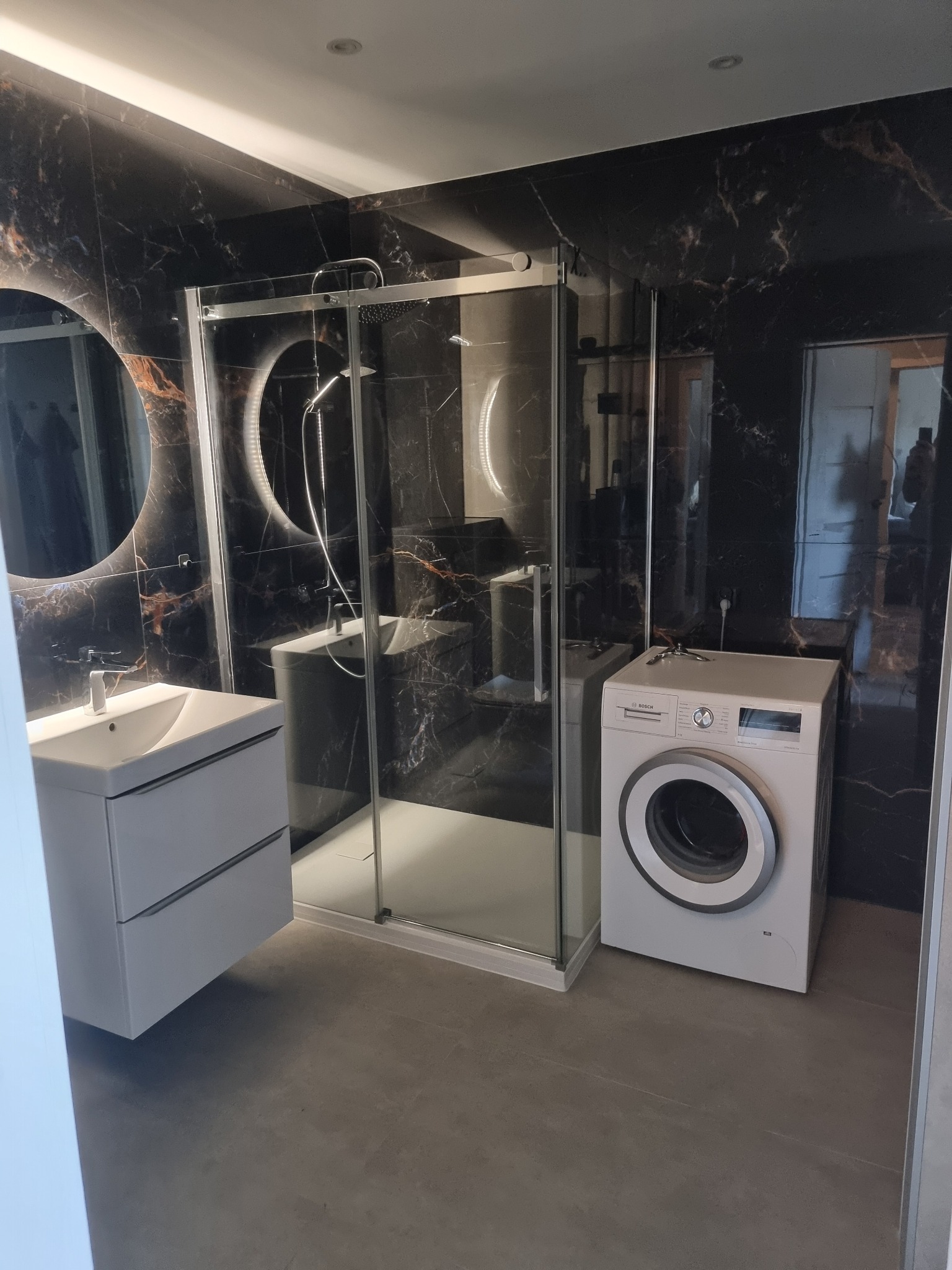 Nowoczesna łazienka z czarnymi płytkami imitującymi marmur, kabiną prysznicową, białą umywalką z szafką i pralką. Okrągłe lustro z podświetleniem.