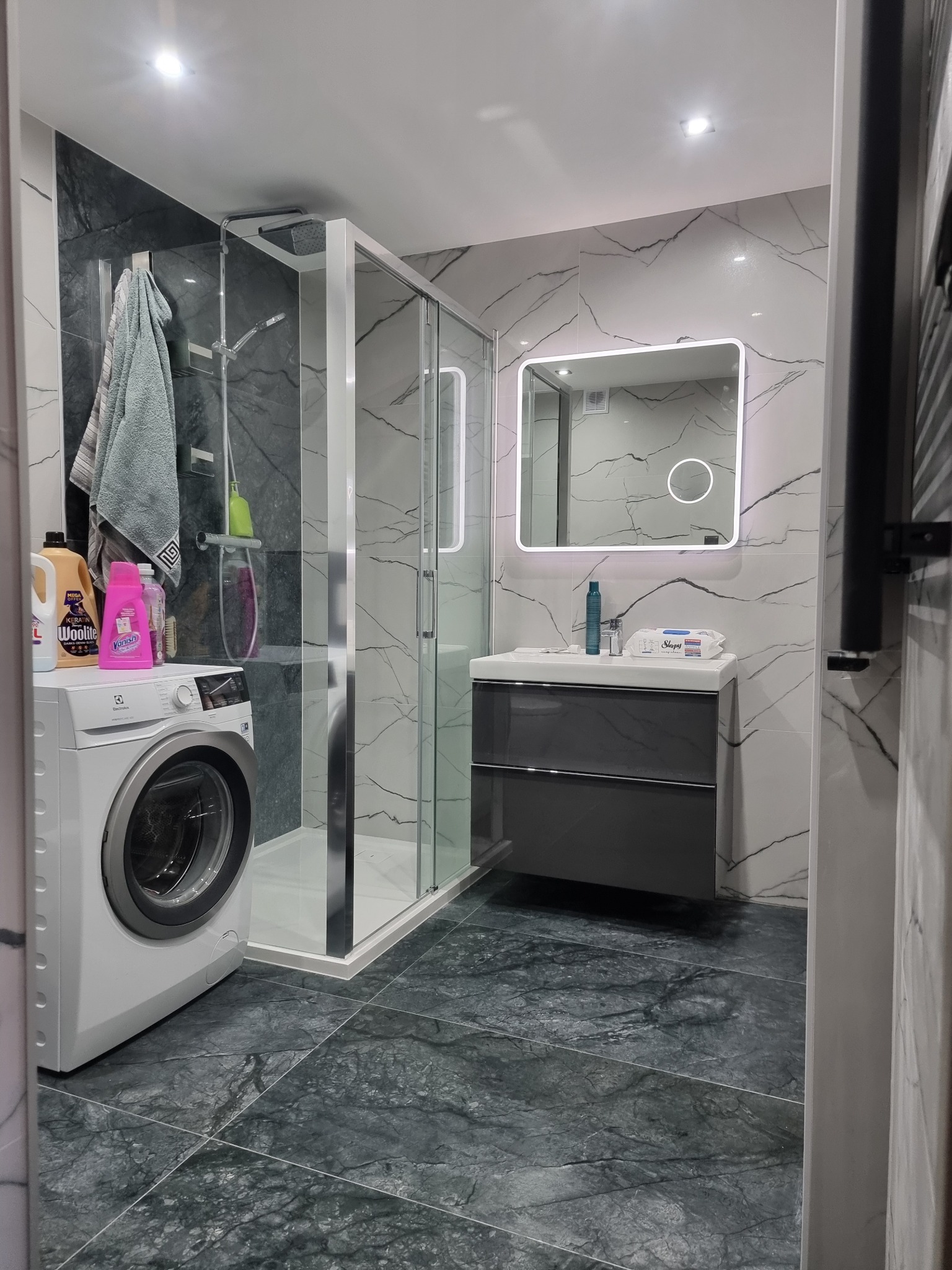 Nowoczesna łazienka po remoncie w Świnoujściu: kabina prysznicowa, umywalka z szafką, pralka. Marmurowe płytki na ścianach i podłodze. Oświetlone lustro.