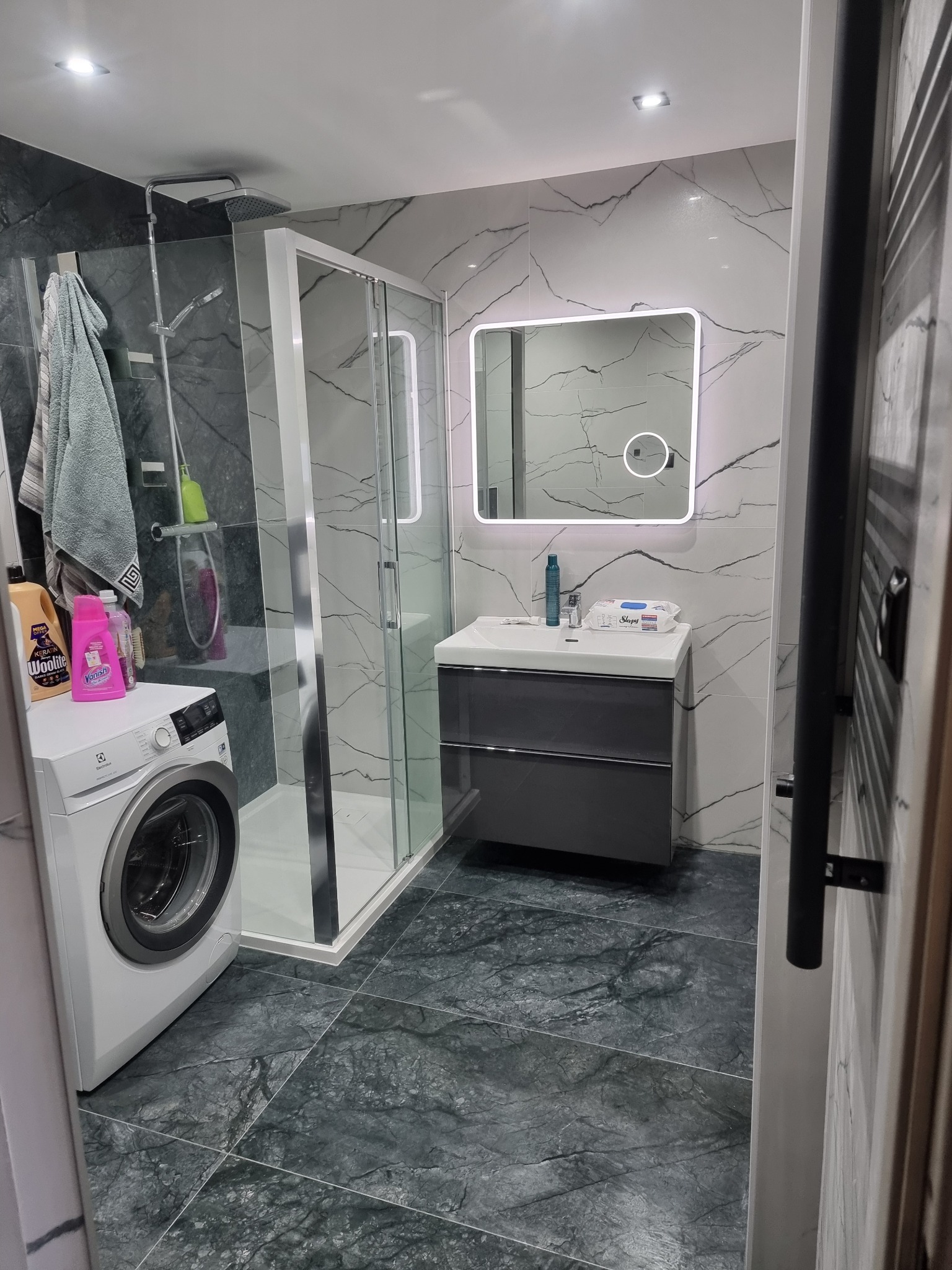 Nowoczesna łazienka po remoncie: kabina prysznicowa, umywalka z szafką, pralka i marmurowe płytki na ścianach i podłodze. Całość utrzymana w szaro-białej kolorystyce.