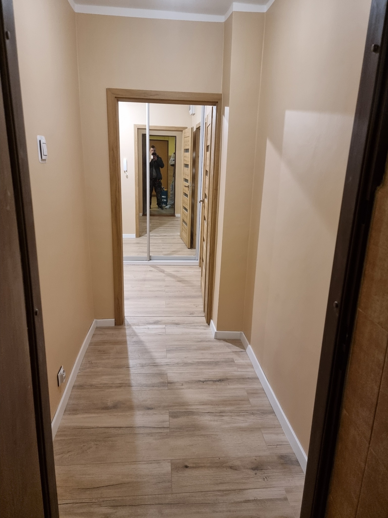 Beżowy korytarz z drewnopodobną podłogą i lustrem w zabudowie. Widoczna osoba robiąca zdjęcie. Minimalistyczne wykończenie, ciepłe oświetlenie.