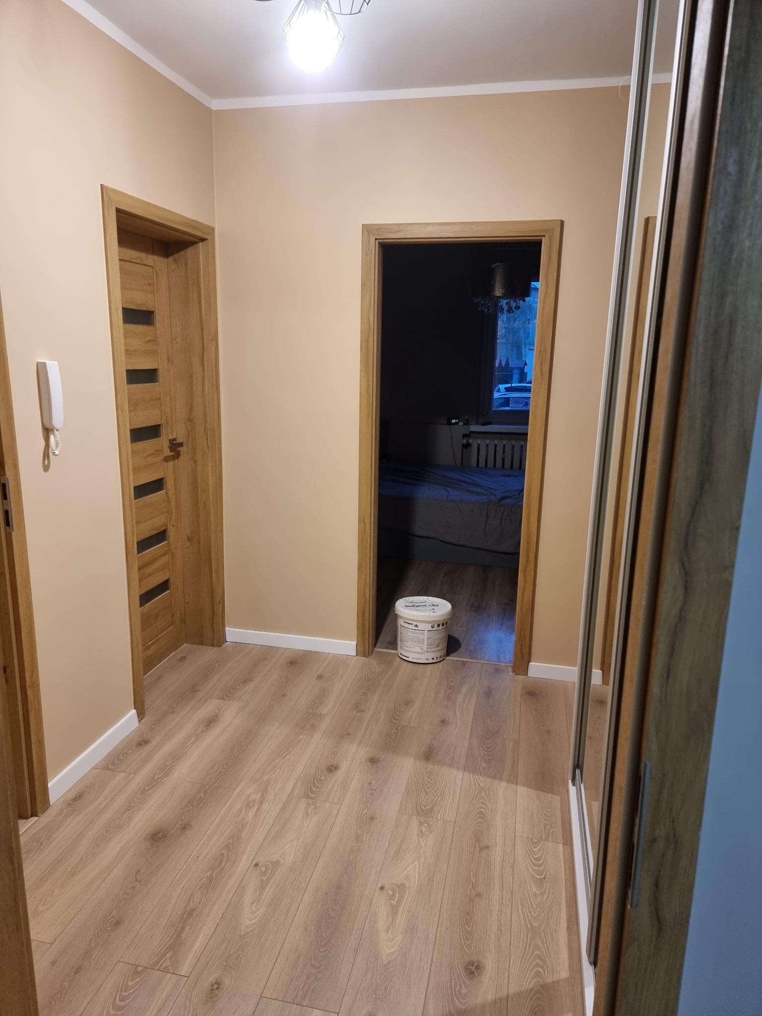 Beżowy korytarz po remoncie z drewnianą podłogą, dwojgiem drzwi i białą puszką farby. Widoczna szafa z lustrem i lampa sufitowa.