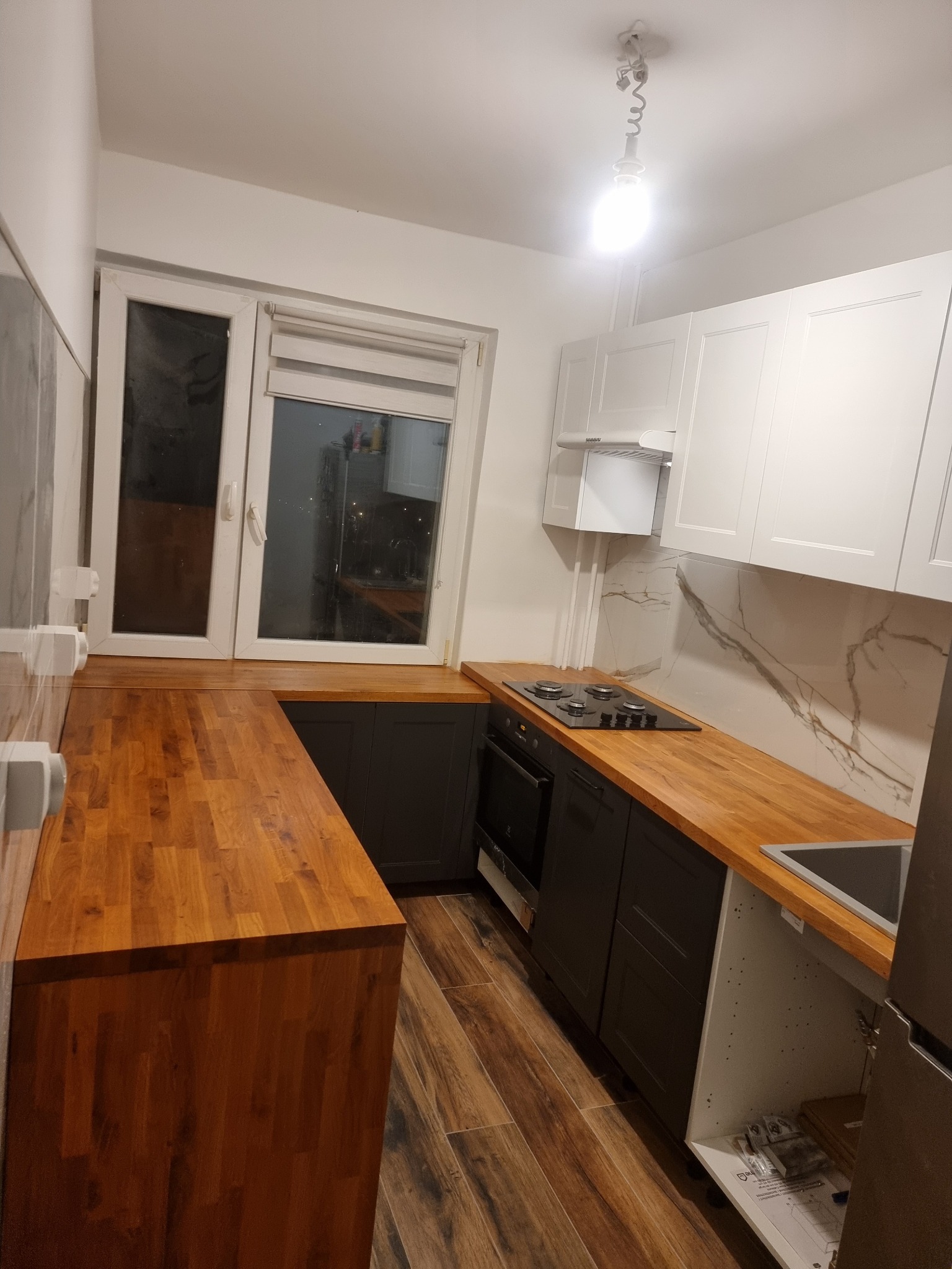 Nowoczesna kuchnia w bloku: drewniany blat, szare szafki dolne, białe górne, marmurowe wykończenie ściany. Widok na okno i fragment podłogi z paneli.