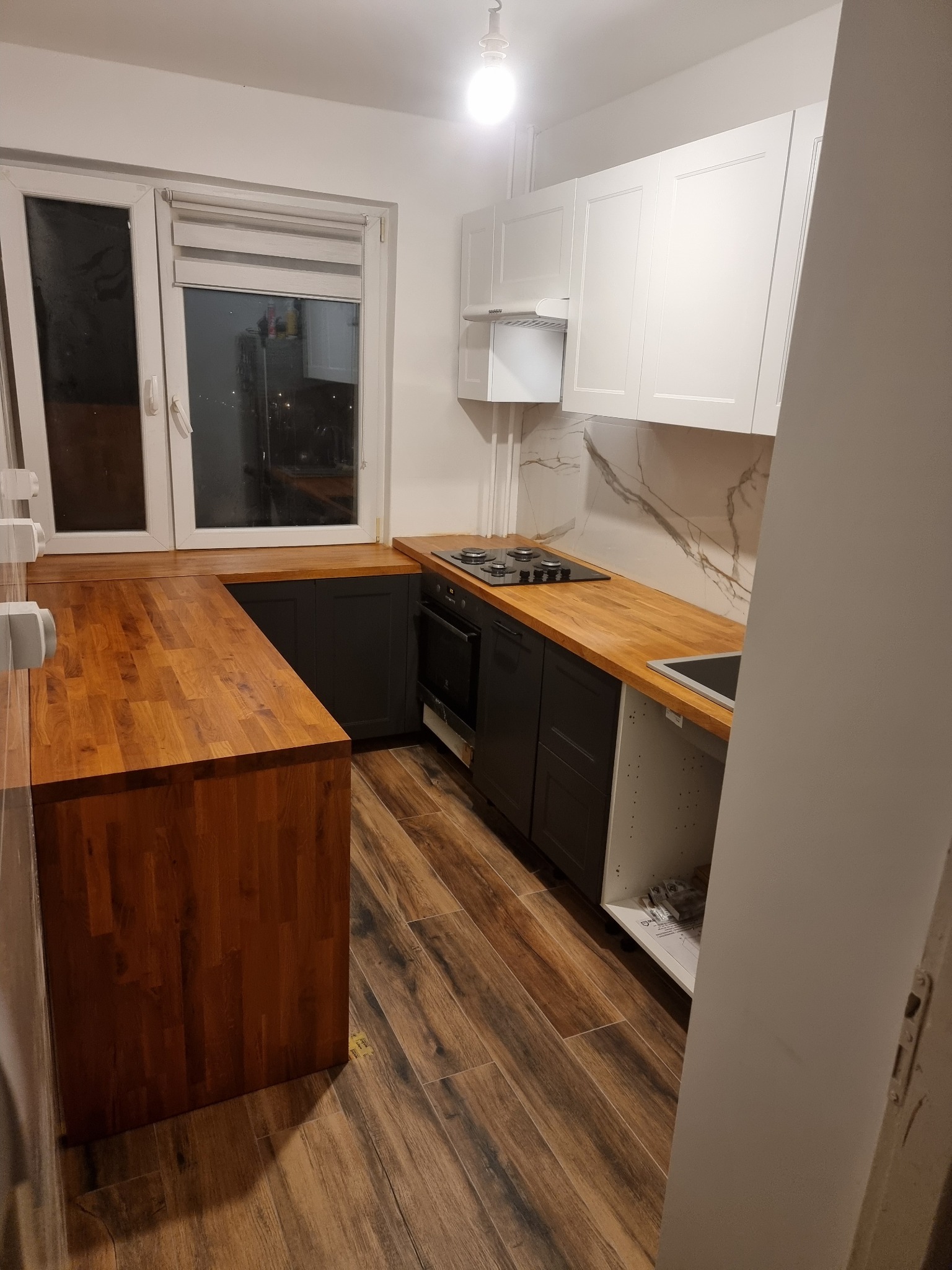 Nowoczesna kuchnia po remoncie: drewniany blat, ciemne szafki dolne, białe górne, marmurowe wykończenie ściany nad blatem, drewnopodobna podłoga.