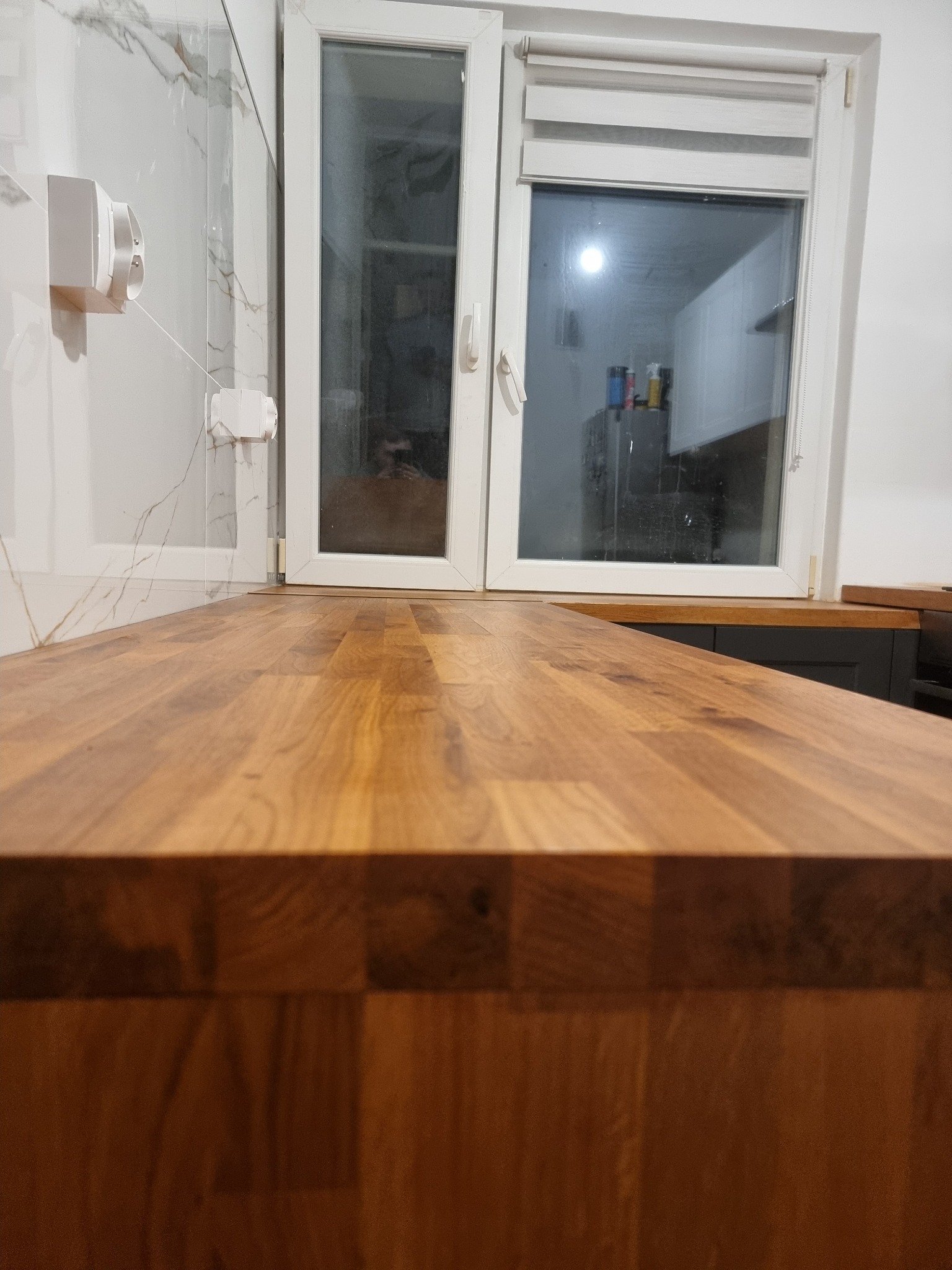 Drewniany blat kuchenny z widocznym oknem i marmurową ścianą w tle. Ujęcie z niskiej perspektywy podkreśla strukturę drewna i nowoczesny design kuchni.