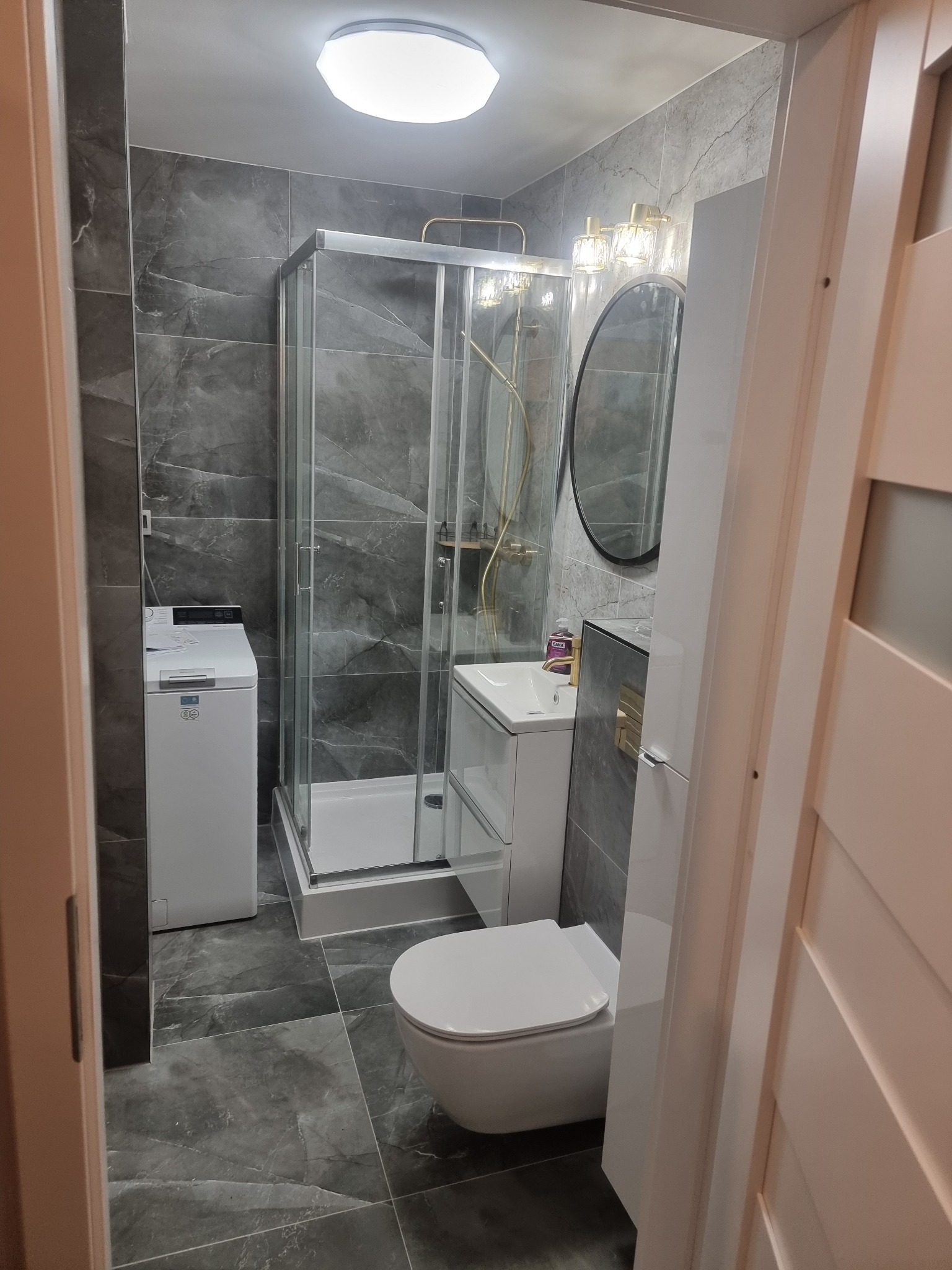 Wykończona łazienka w szarościach: kabina prysznicowa, umywalka z szafką, toaleta i pralka. Marmurowe płytki na ścianach i podłodze, oświetlenie sufitowe i nad lustrem.