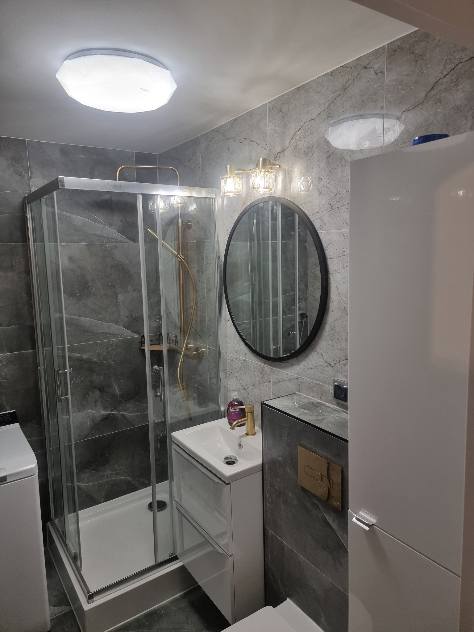 Nowoczesna łazienka z kabiną prysznicową, umywalką z szafką, okrągłym lustrem i szarymi płytkami imitującymi marmur. Złote akcenty armatury i oświetlenia.