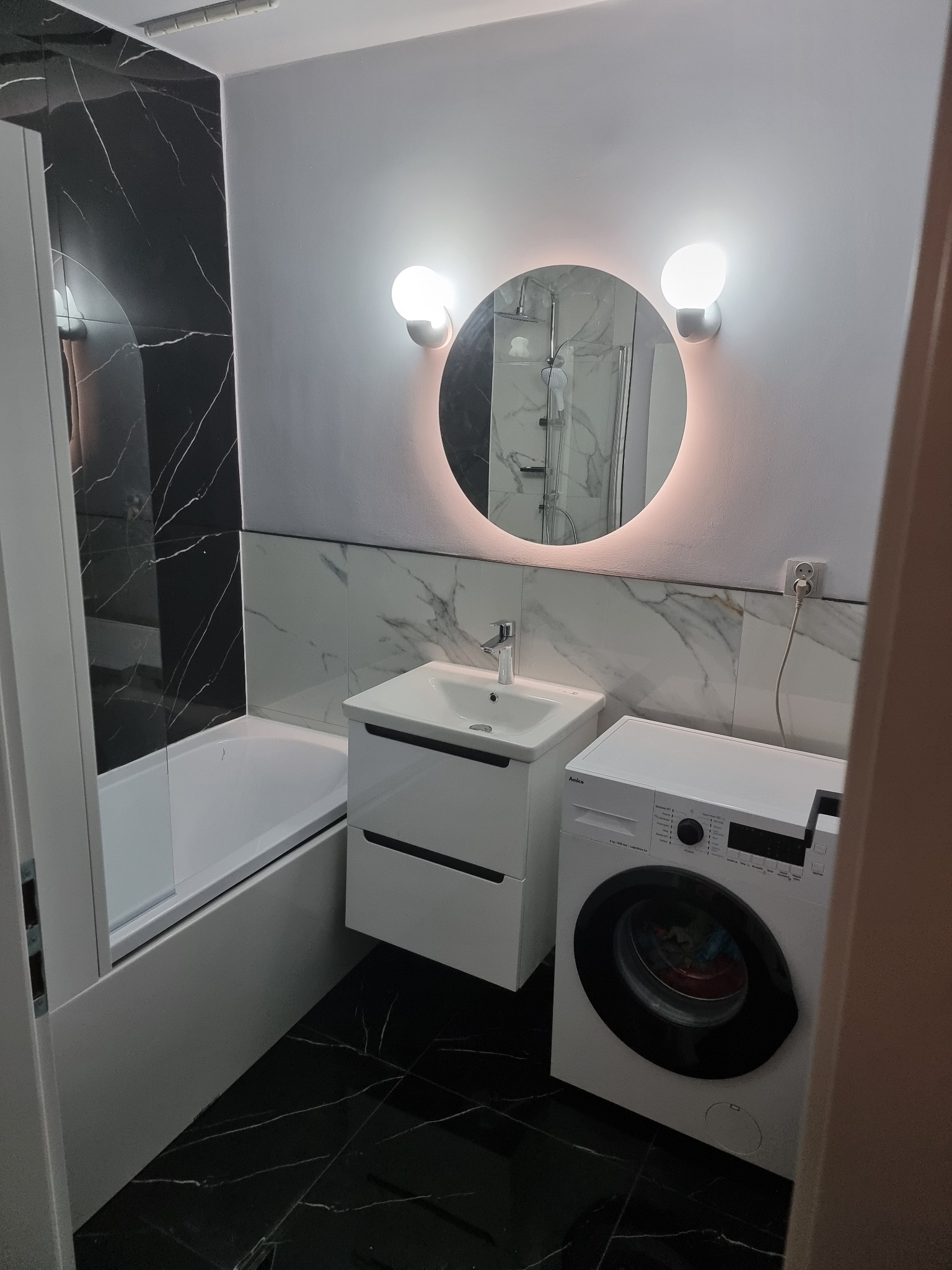 Nowoczesna łazienka po remoncie: czarne płytki imitujące marmur, okrągłe lustro z podświetleniem LED, biała umywalka z szafką i pralka. Minimalistyczny design.