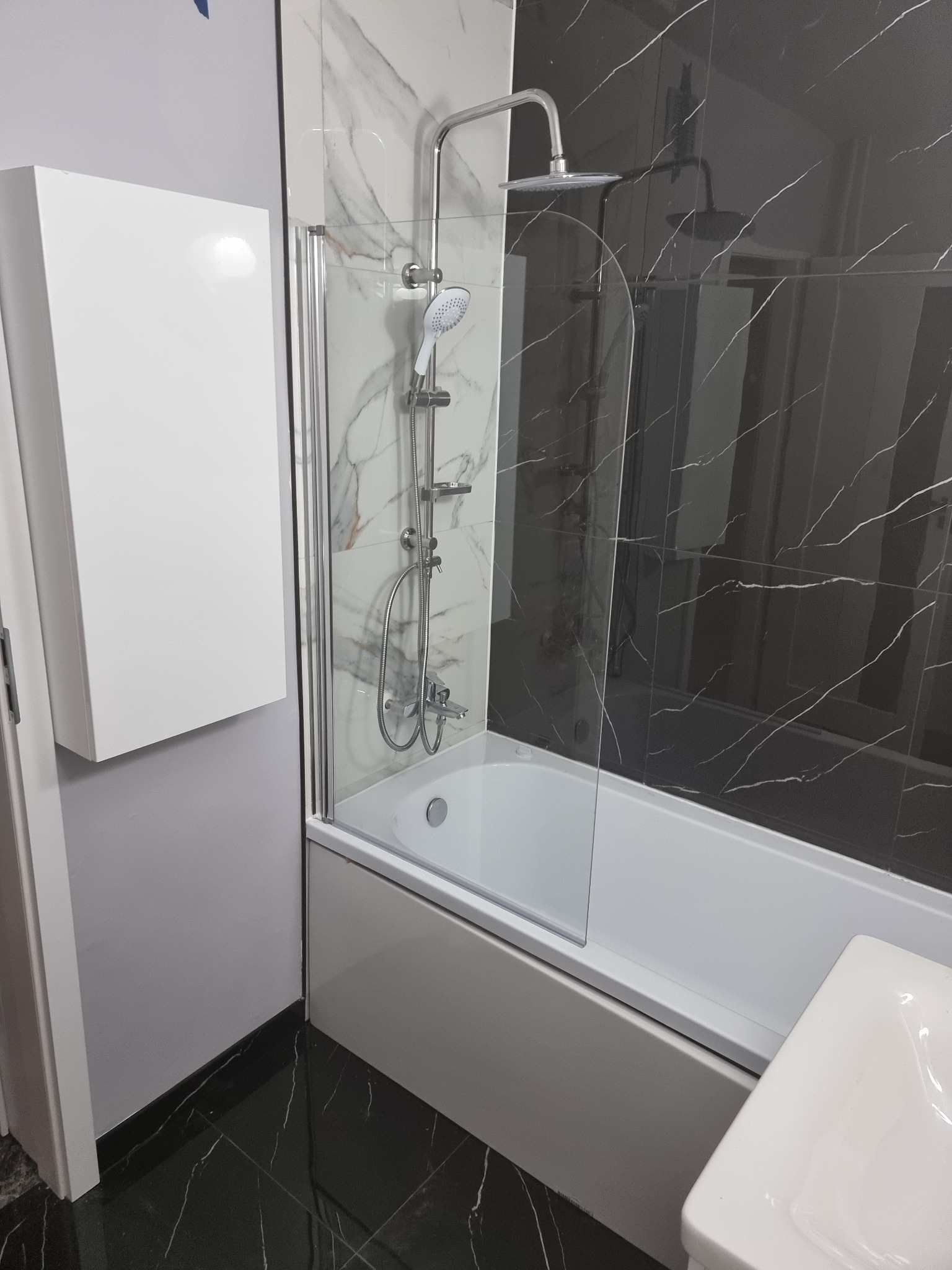 Nowoczesna łazienka z czarno-białymi marmurkowymi płytkami, białą wanną z parawanem i szafką. Czarne płytki podłogowe z białymi żyłkami.