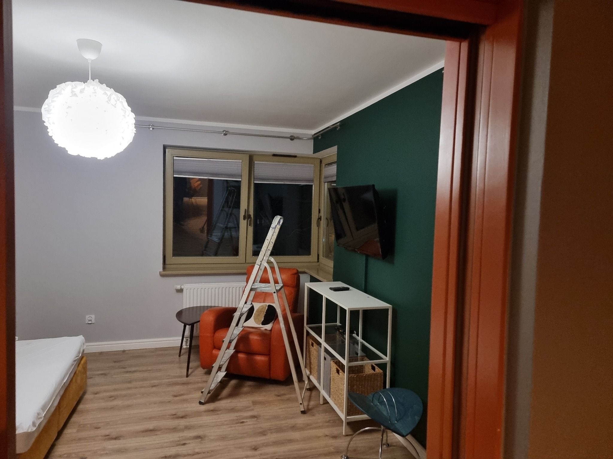 Pokój po odświeżeniu: szara ściana, zielona ściana z TV, drabina oparta o fotel, biała lampa, drewniana podłoga. Widok zza framugi drzwi.