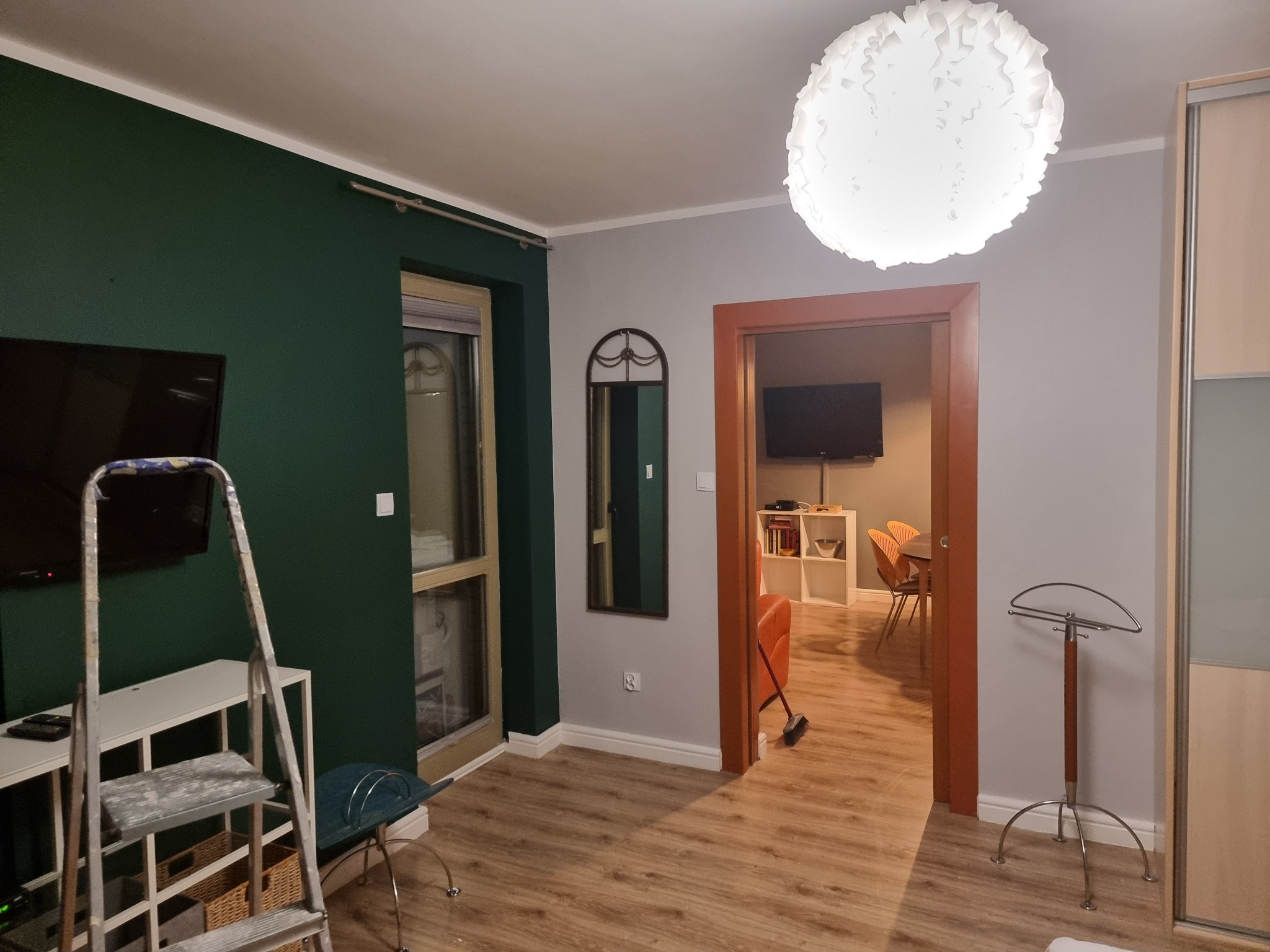 Wnętrze pokoju po malowaniu ścian na zielono i szaro, widoczna drabina malarska, lustro na ścianie i lampa sufitowa. Podłoga wyłożona panelami.