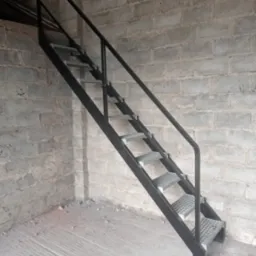Metalowe schody drabinkowe z poręczą, prowadzące w górę, ustawione przy ścianie z szarej cegły