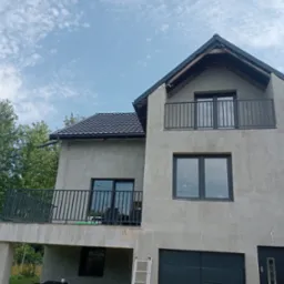 Dom jednorodzinny z szarym tynkiem i czarnymi balustradami balkonowymi. Widoczna drabina oparta o ścianę. Ujęcie z dołu, na tle błękitnego nieba.