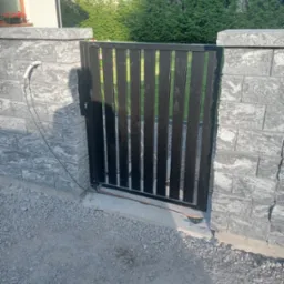 Nowoczesna, czarna furtka z pionowych profili, osadzona między szarymi, betonowymi słupkami. Widoczne okablowanie przy jednym ze słupk&oacute;w.