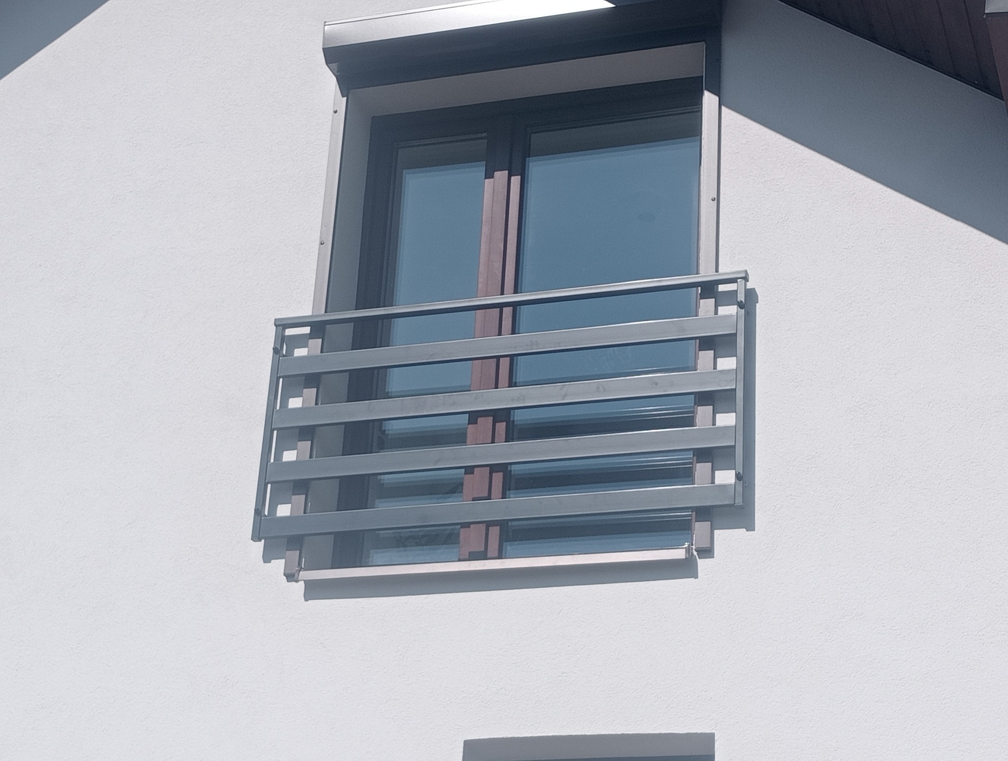 Szara, pozioma balustrada okienna z prostych, metalowych prętów na tle białej fasady budynku.