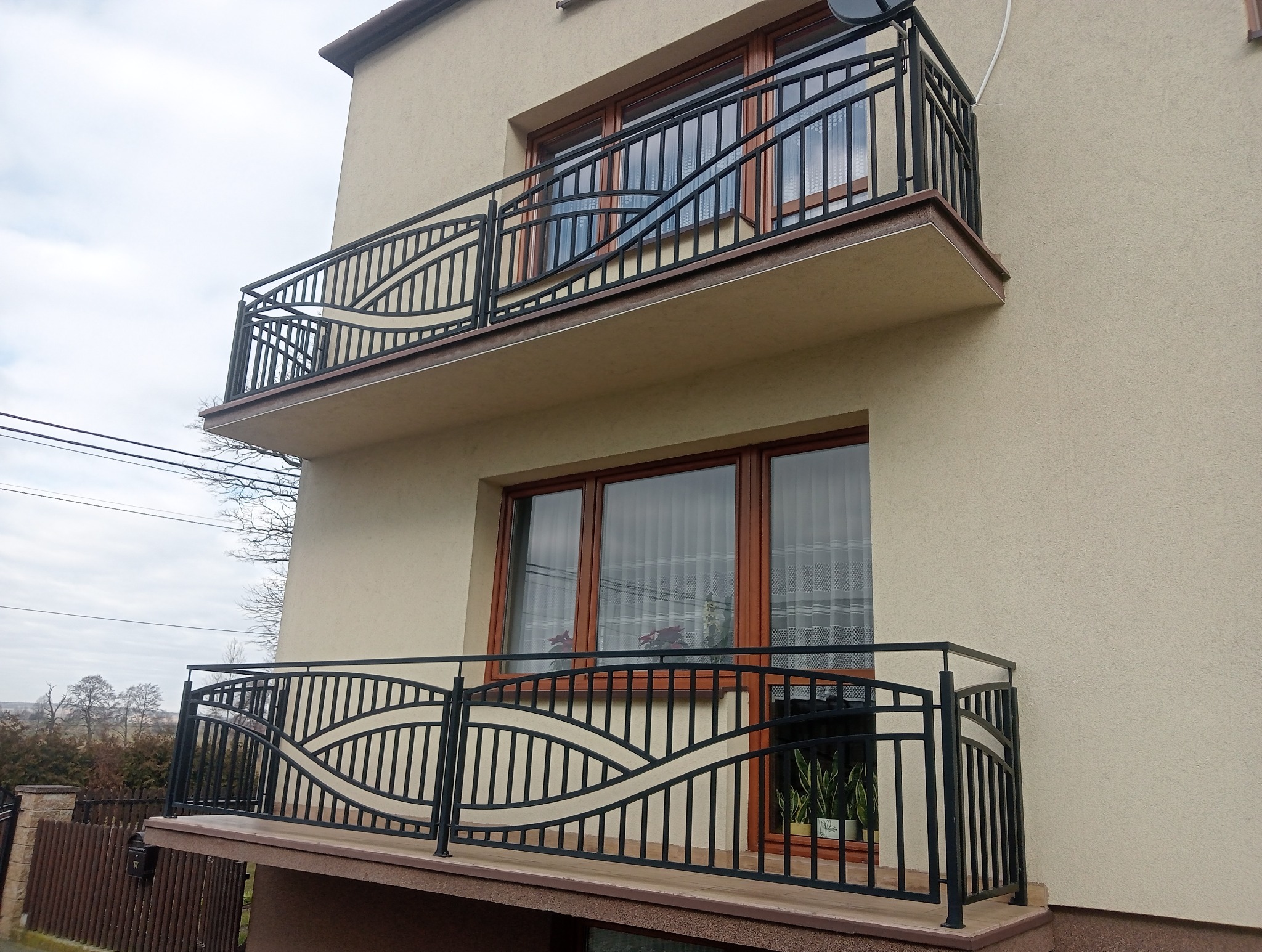 Dwa balkony z czarnymi, metalowymi balustradami o dekoracyjnym, falistym wzorze, zamontowane na beżowej elewacji budynku z brązowymi ramami okiennymi.