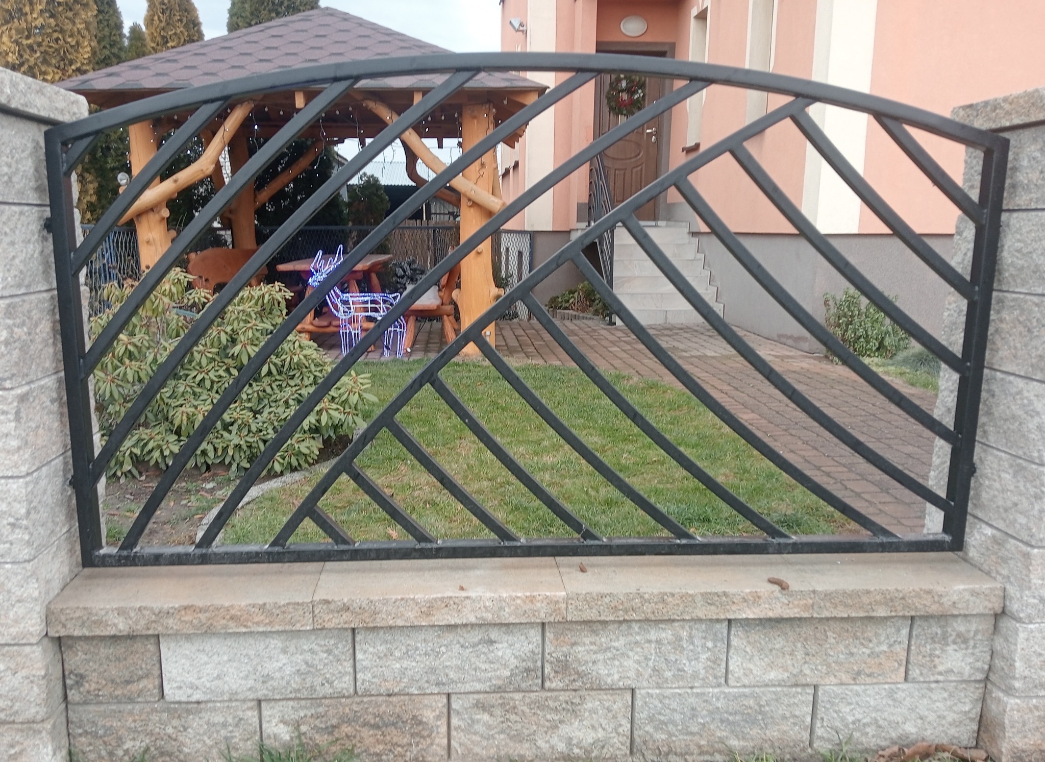Fragment czarnego ogrodzenia z metalowych prętów na niskim murku z kamieni, w tle trawnik, altana i budynek mieszkalny.