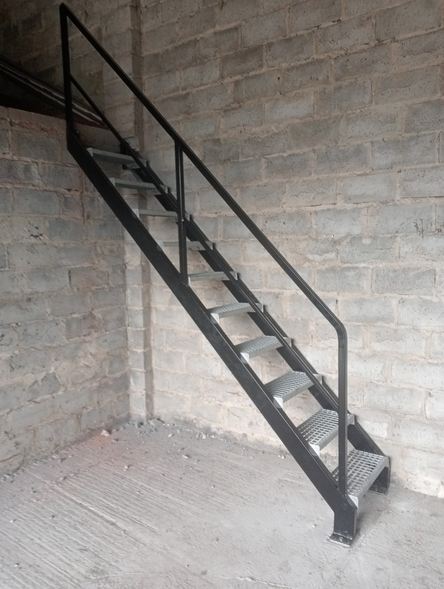 Metalowe schody drabinkowe z poręczą, prowadzące w górę, ustawione przy ścianie z szarej cegły