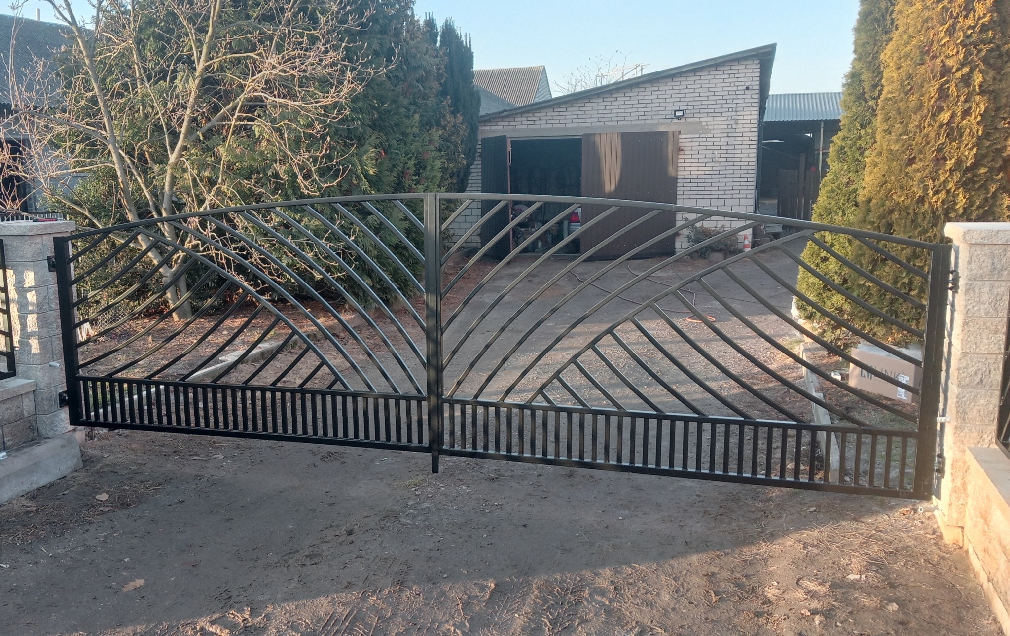 Czarna, metalowa brama wjazdowa z dekoracyjnym wzorem wachlarza na tle posesji z garażem i zielenią