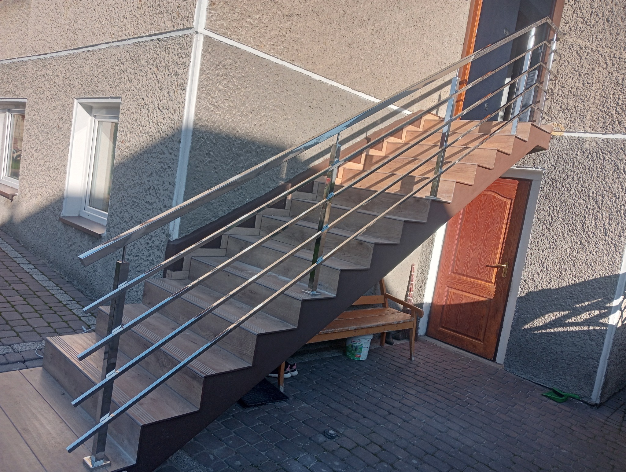 Zewnętrzne schody z okładziną imitującą drewno, z balustradą ze stali nierdzewnej, prowadzące do drzwi wejściowych budynku o szarej, chropowatej elewacji.