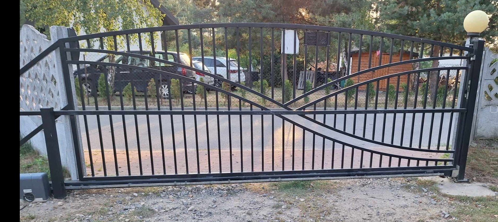 Czarna, przesuwna brama wjazdowa z metalowymi prętami i ozdobnymi łukami, widoczny automat do bramy, oświetlenie słupkowe i częściowo samochody w tle.