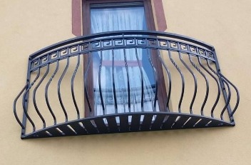 Kuty balkon z czarną, giętą balustradą o motywie greckim, zamontowany na żółtej elewacji budynku z oknem w tle.