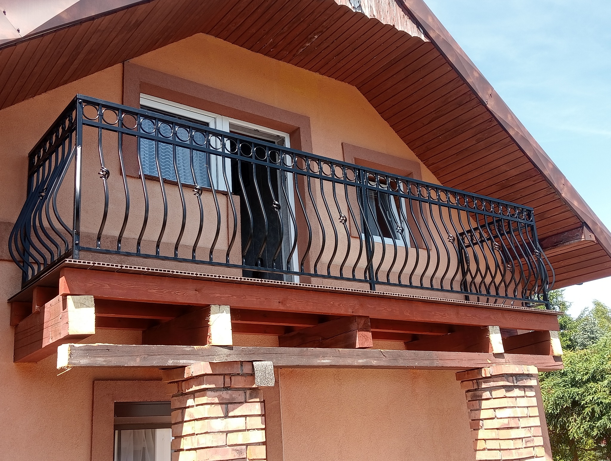 Czarna, kuta balustrada balkonowa z dekoracyjnymi elementami na tle jasnej elewacji domu z drewnianym zadaszeniem.