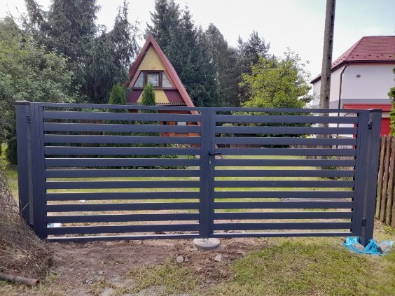 Nowoczesne, poziome przęsła ogrodzeniowe w kolorze grafitowym, z widocznym zamkiem i słupkami, na tle zieleni i domów.