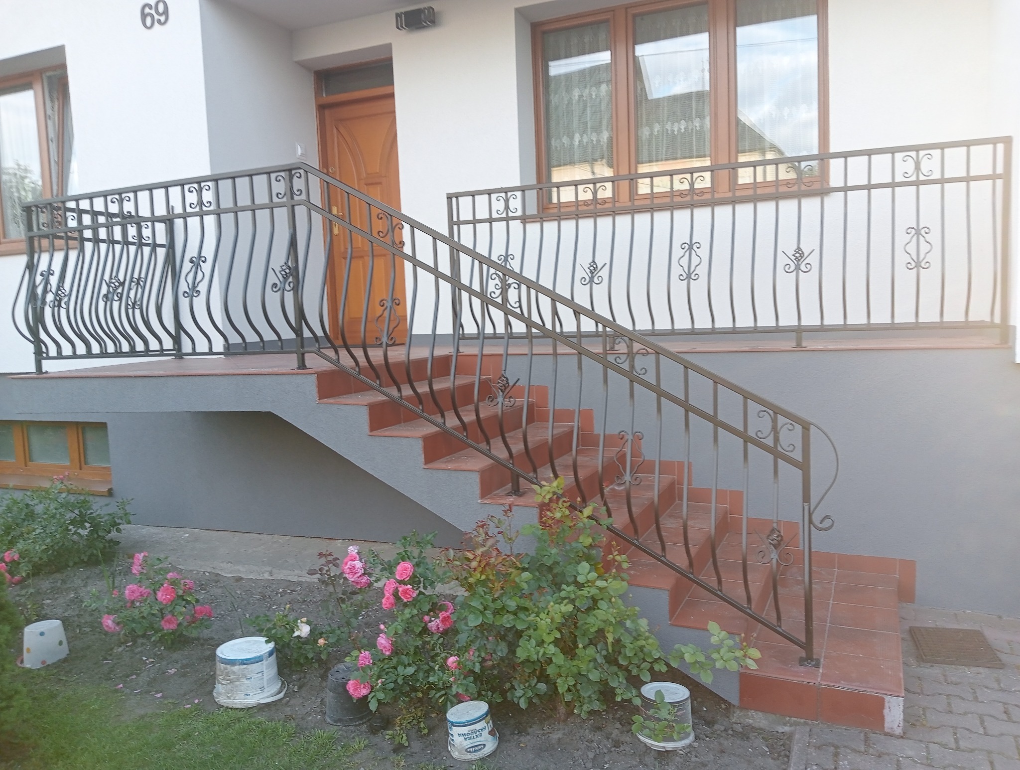 Kute, czarne balustrady schodowe z ozdobnymi elementami, prowadzące do drzwi wejściowych domu z numerem 69, otoczone rabatą róż.