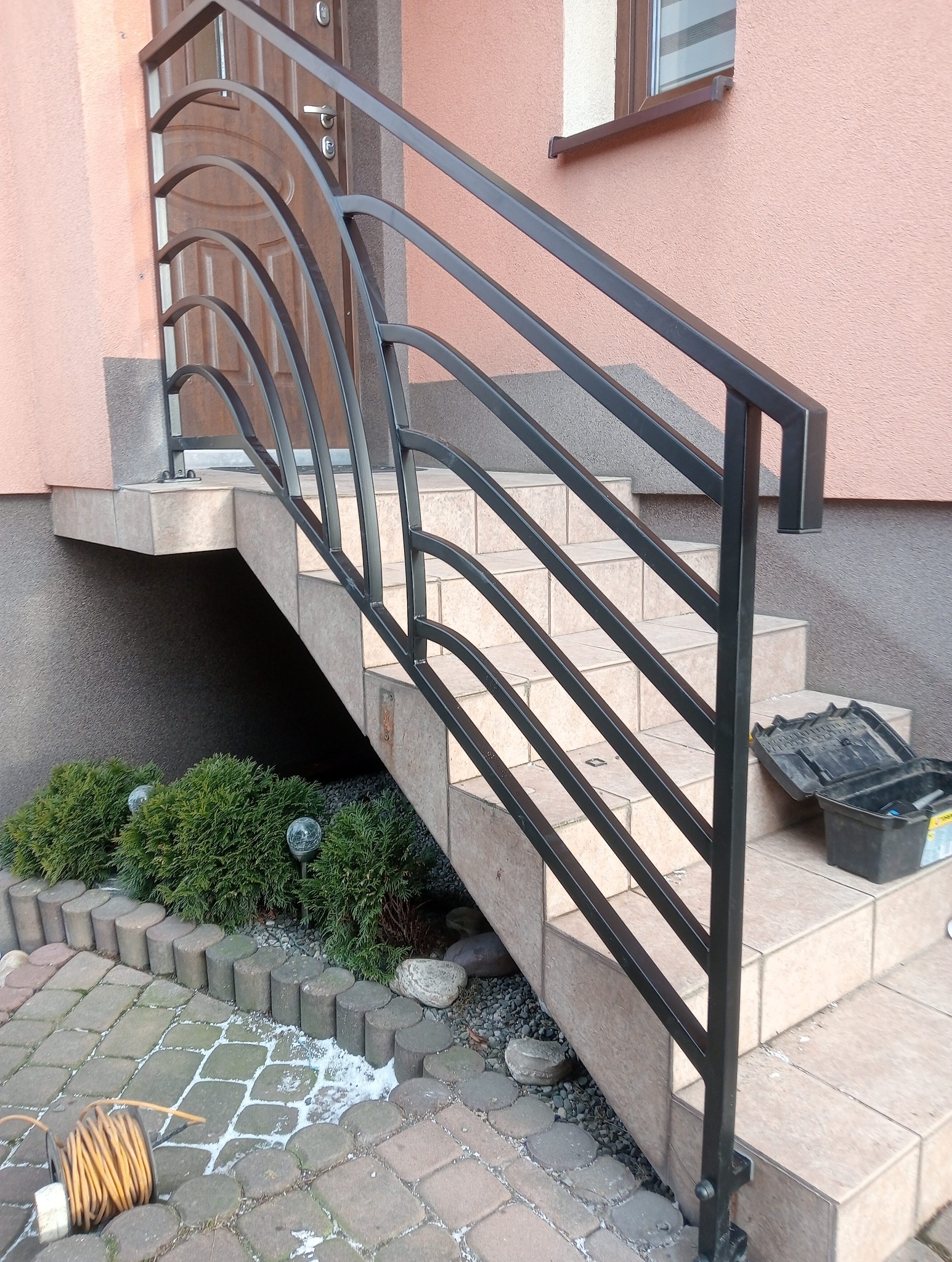 Czarna, metalowa balustrada schodowa z motywem łuków, prowadząca do drzwi wejściowych domu; obok skrzynka z narzędziami i zwinięty kabel elektryczny.