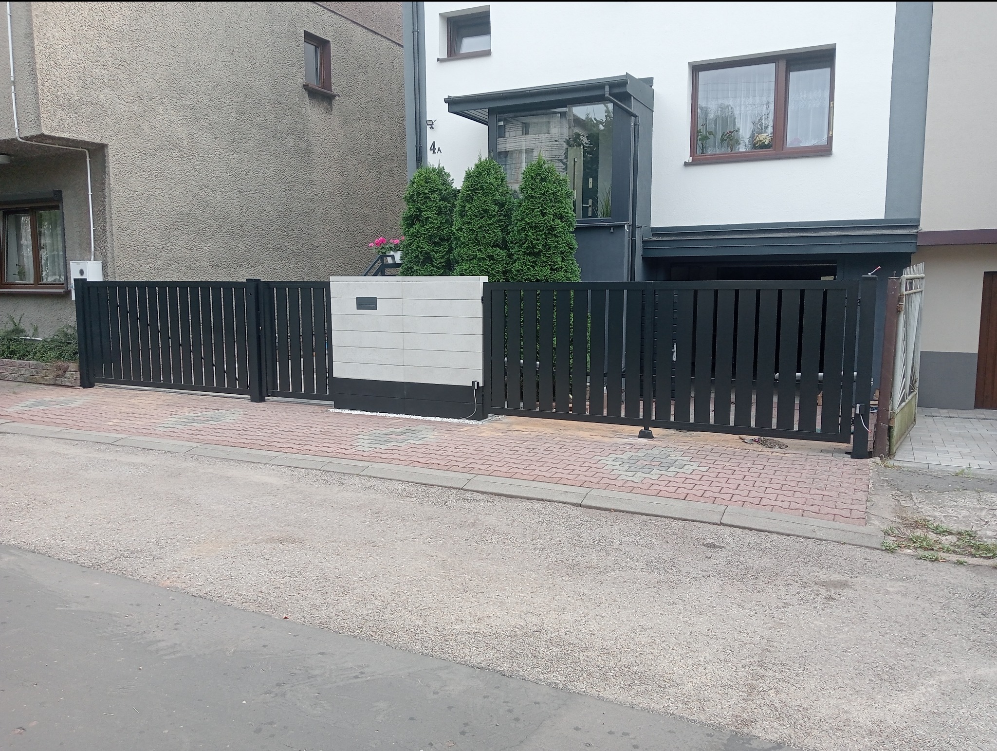 Nowoczesne, grafitowe ogrodzenie panelowe z automatyczną bramą wjazdową, z jasnoszarym, segmentowym słupkiem na tle dwukondygnacyjnego domu.