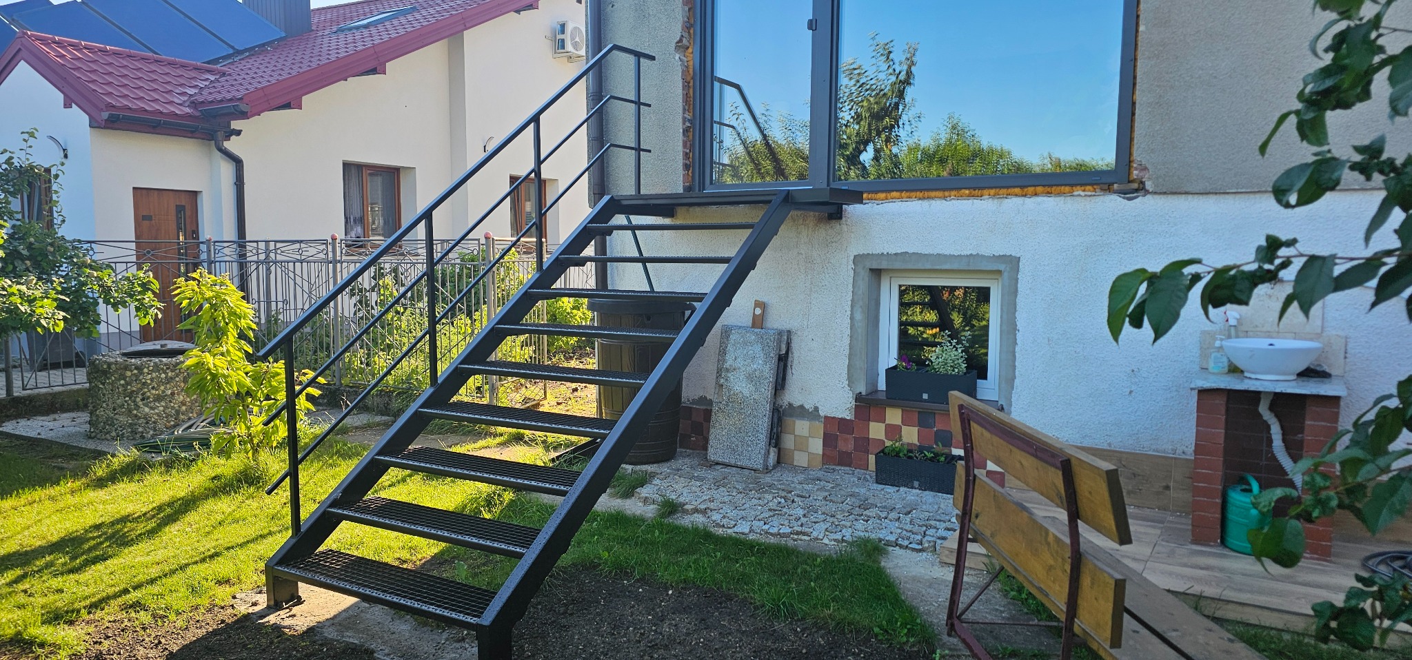 Metalowe schody zewnętrzne prowadzące do okna na piętrze, z czarnymi, ażurowymi stopniami i balustradą, na tle białego domu z czerwonym dachem i zielenią ogrodu.