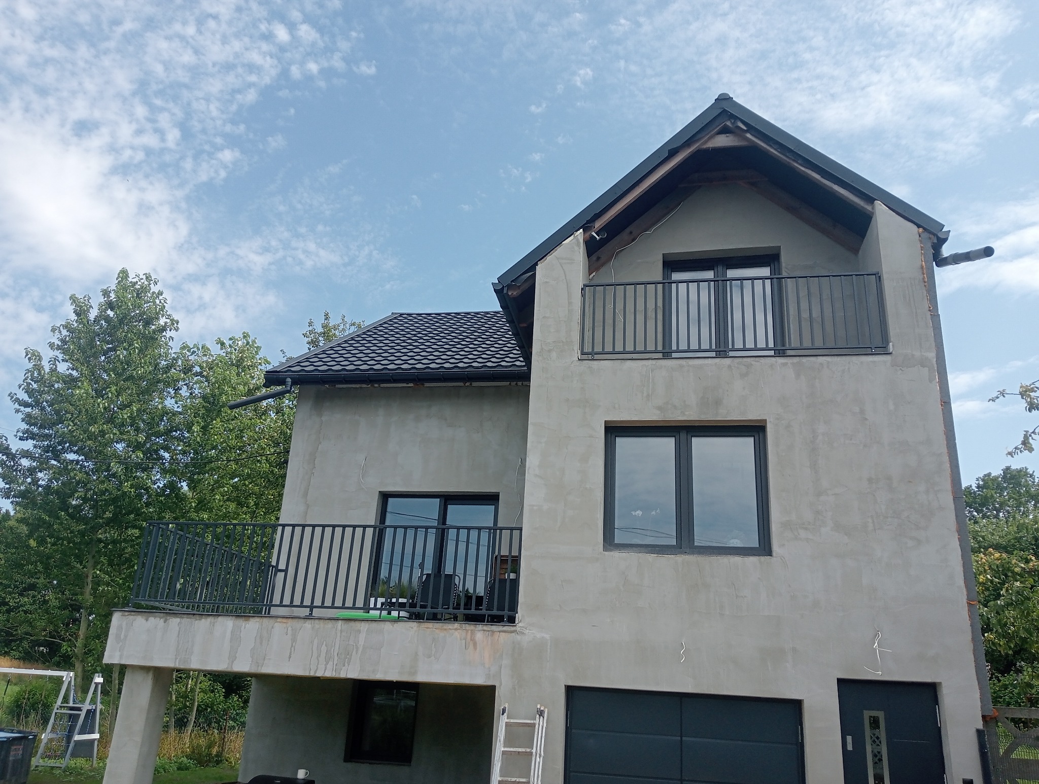 Dom jednorodzinny z szarym tynkiem i czarnymi balustradami balkonowymi. Widoczna drabina oparta o ścianę. Ujęcie z dołu, na tle błękitnego nieba.