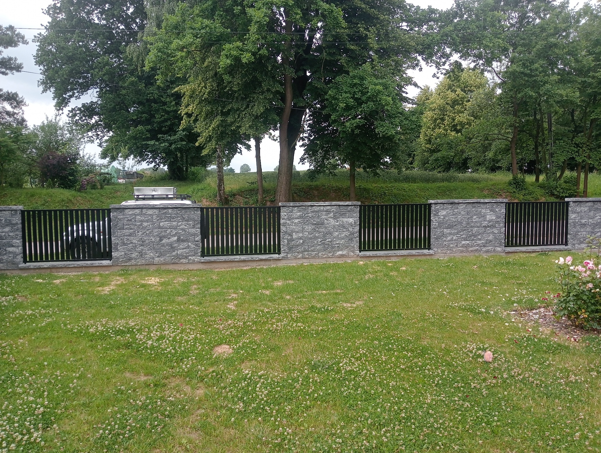 Nowoczesne ogrodzenie metalowe z elementami z szarej cegły, otoczone zielonym trawnikiem i drzewami. Widoczny samochód za ogrodzeniem.