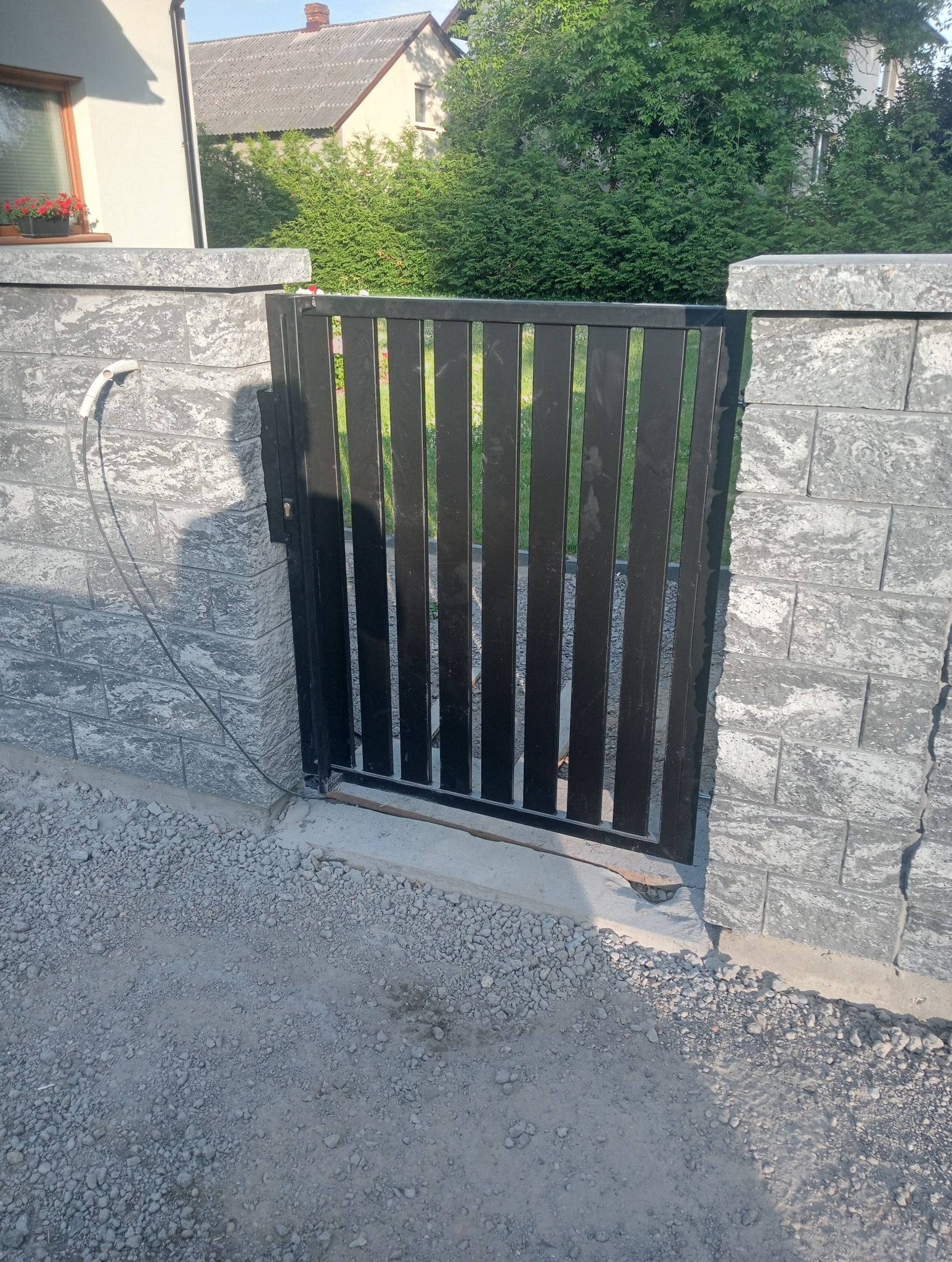 Nowoczesna, czarna furtka z pionowych profili, osadzona między szarymi, betonowymi słupkami. Widoczne okablowanie przy jednym ze słupków.