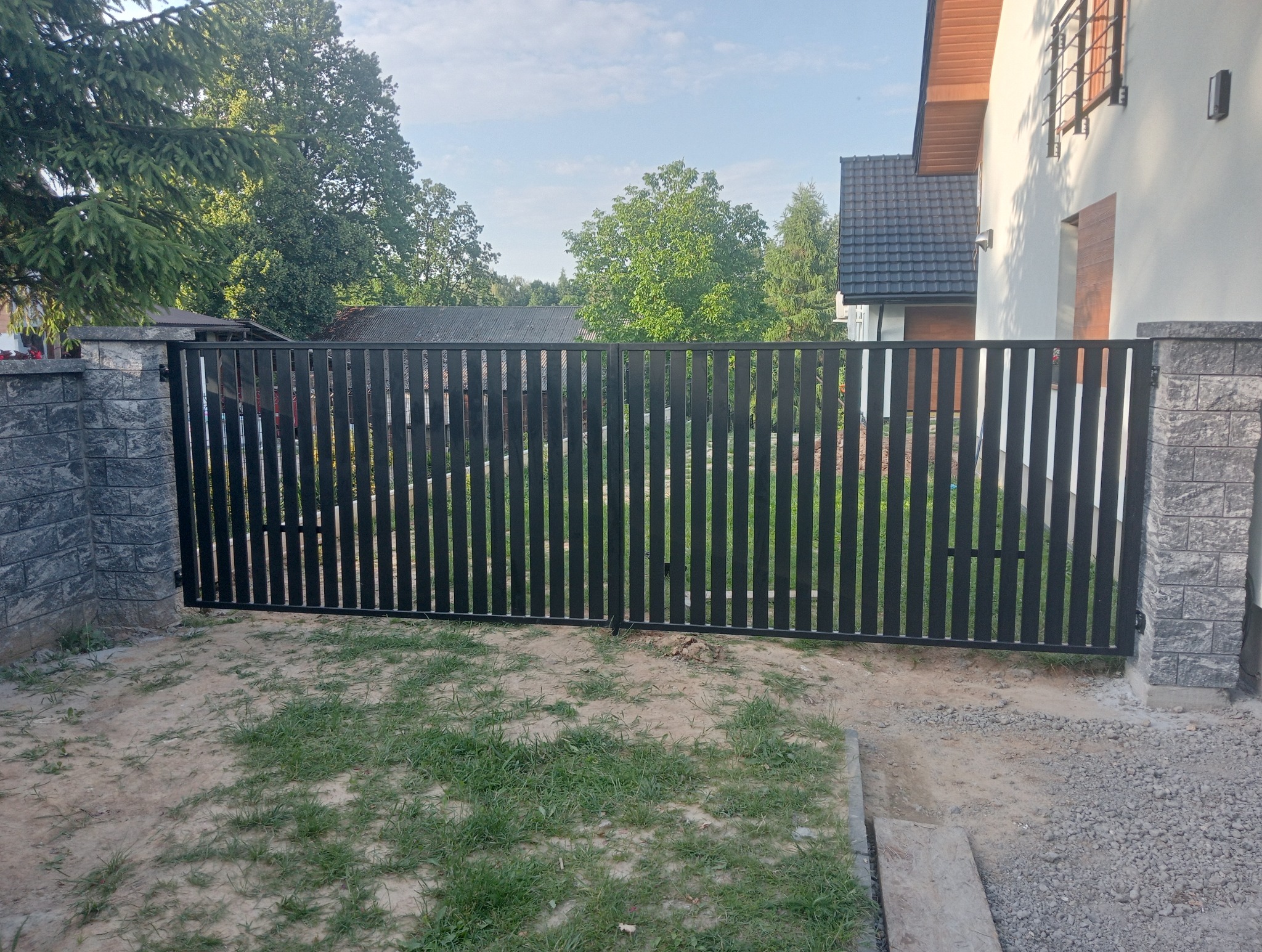 Nowoczesna, metalowa brama wjazdowa z pionowych profili, osadzona na murowanych słupkach z szarej kostki. Tło: dom jednorodzinny i zieleń.