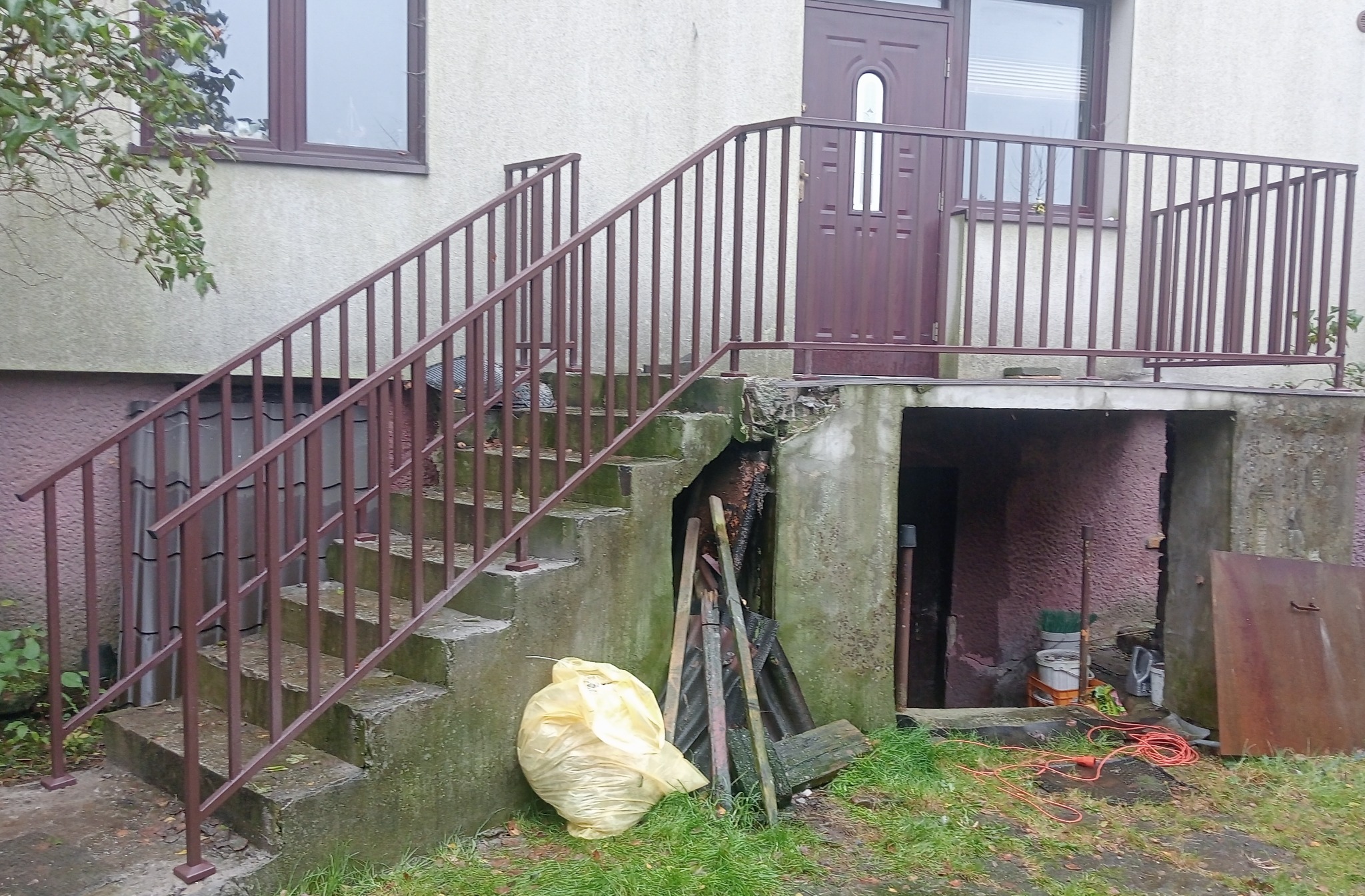 Brązowe, metalowe barierki schodowe i balkonowe przy wejściu do domu. Widoczny fragment elewacji, okno i drzwi wejściowe. Na dole worki i narzędzia.