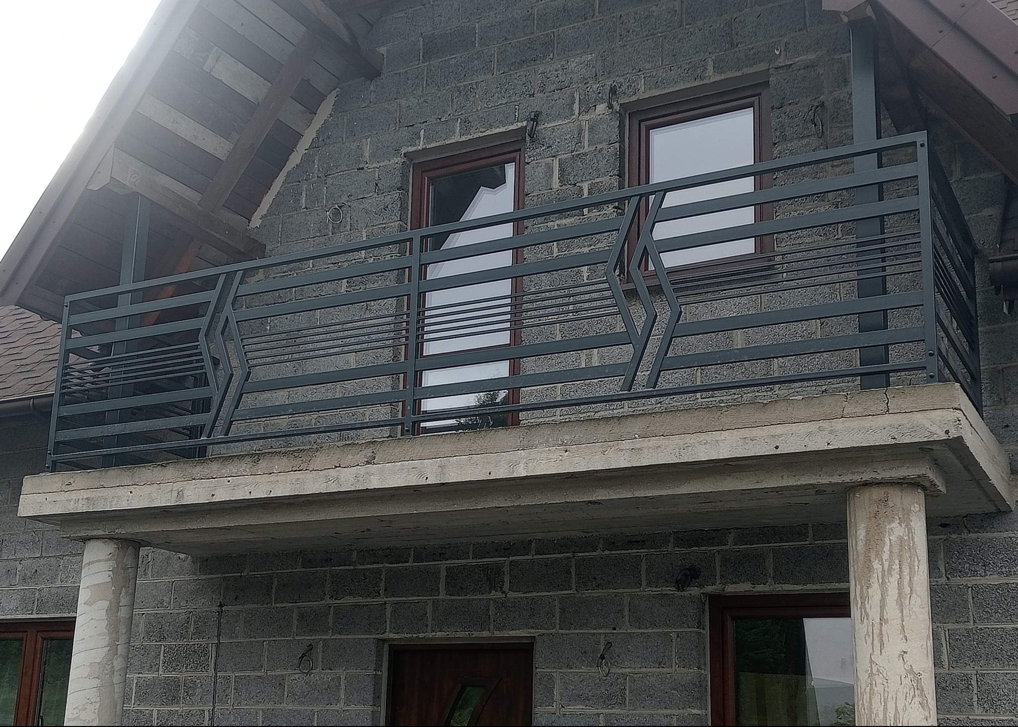 Nowoczesna, pozioma balustrada balkonowa z metalu w kolorze grafitowym, z geometrycznym wzorem, zamontowana na betonowym balkonie domu z szarej cegły.